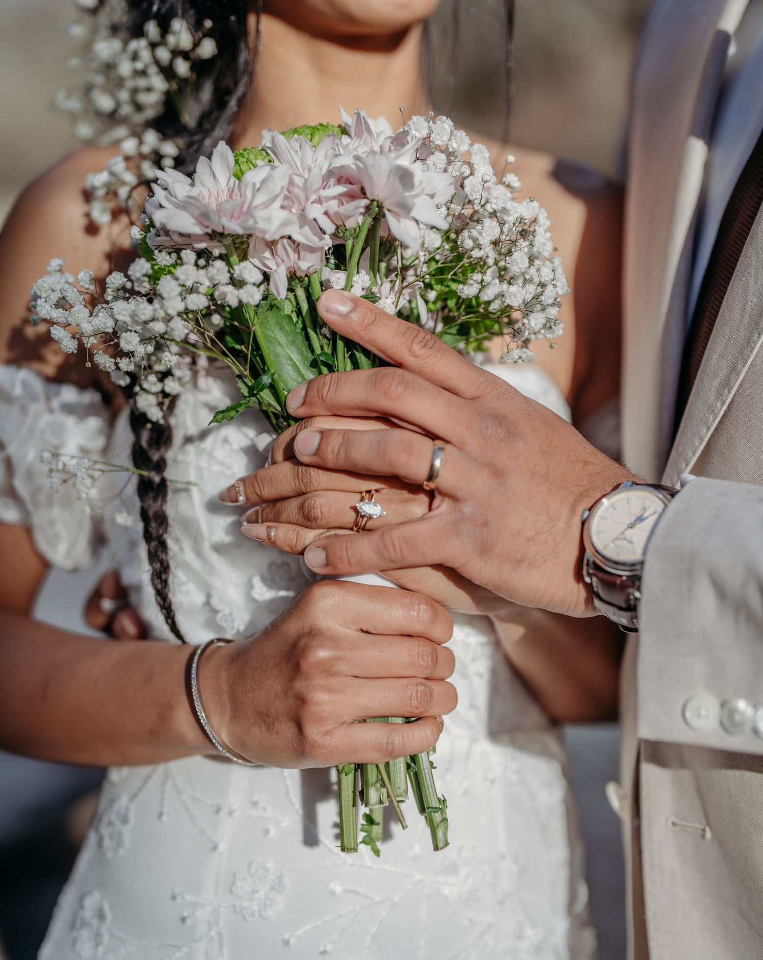 A close-up of wedding rings and hands holding a delicate bouquet of pink chrysanthemums and baby's breath flowers.