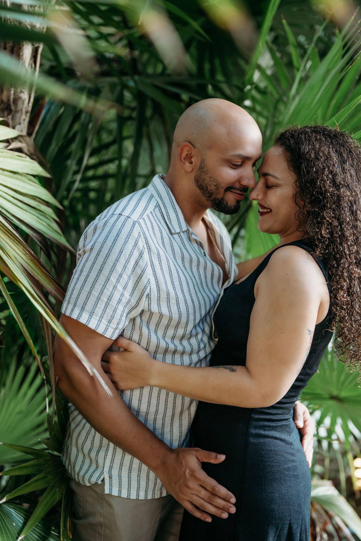 A romantic couple embraces among lush tropical palm leaves during a tender photoshoot moment.