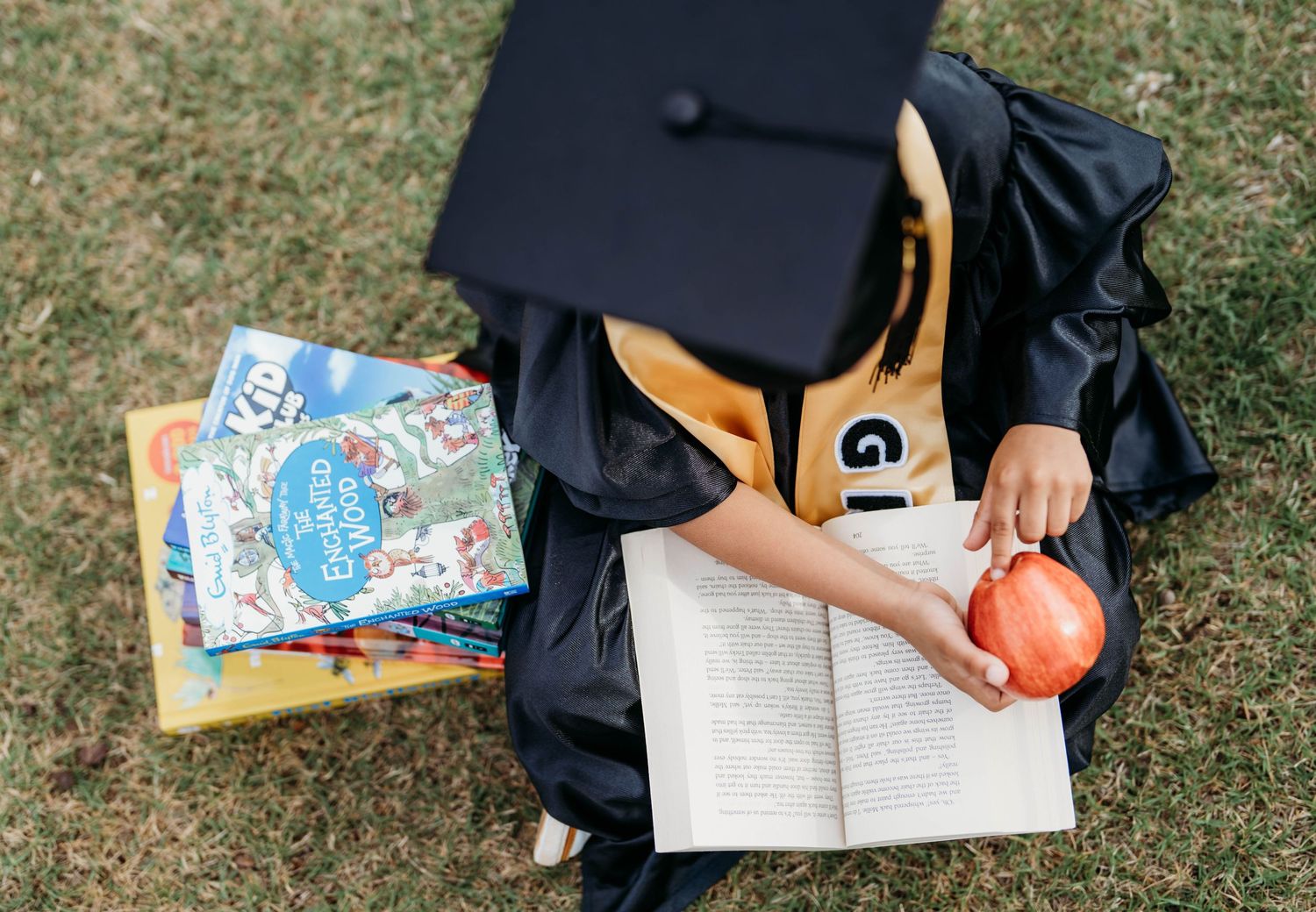 Child in graduation cap and gown reads open book while holding apple, surrounded by colorful childrens books on grass.
