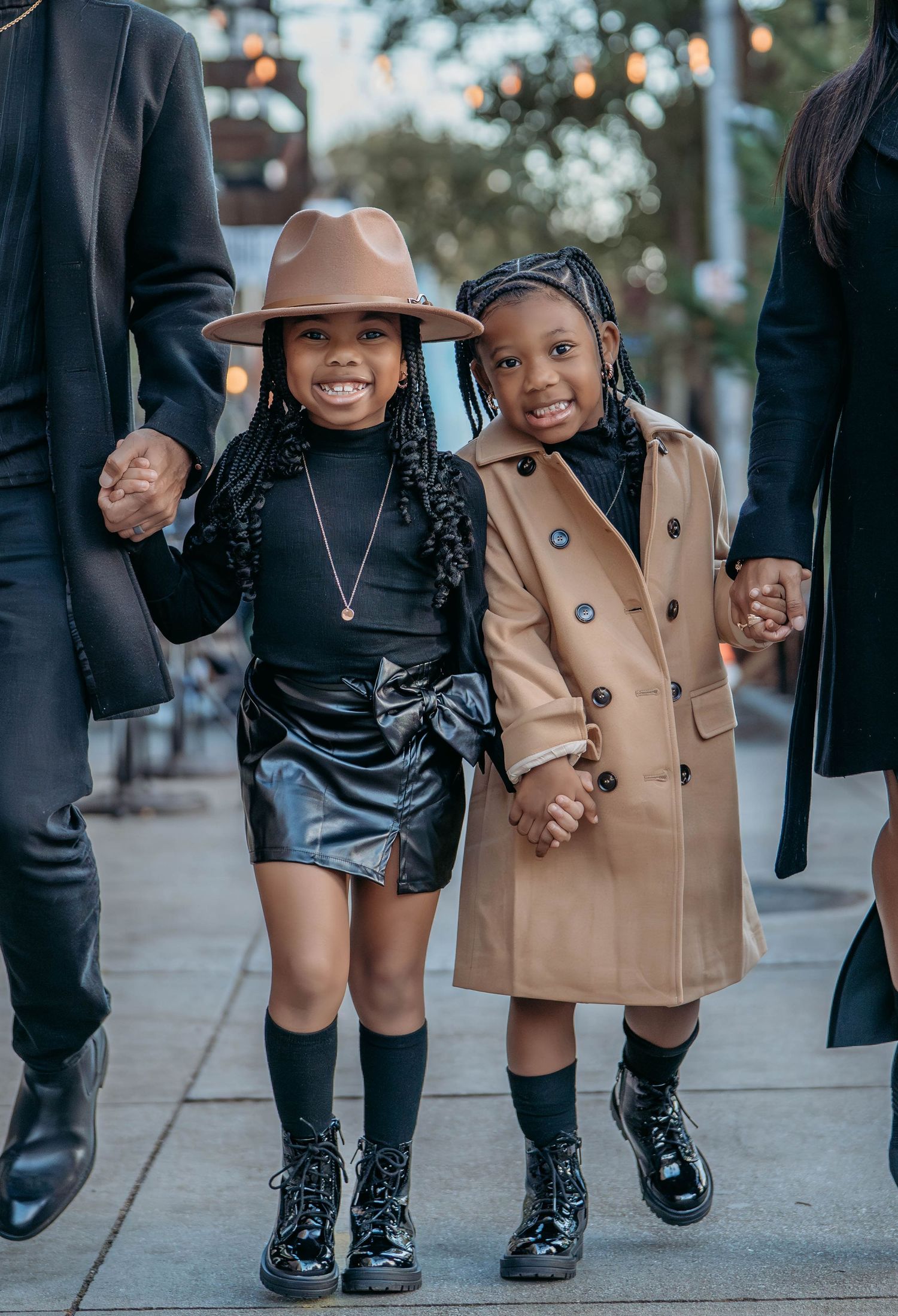 Two children in stylish winter outfits holding hands with adults while walking on a city sidewalk.