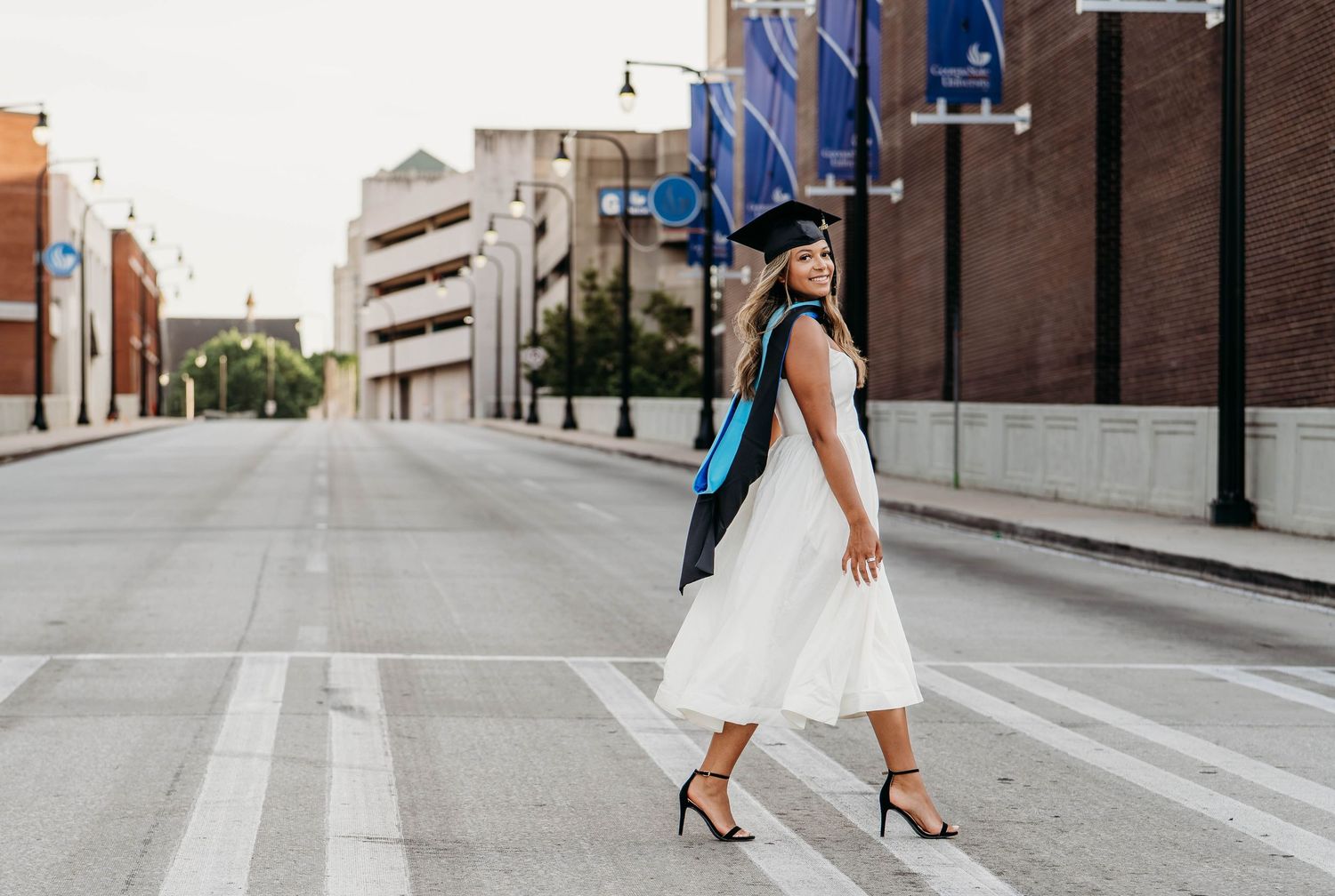 A woman in a white dress and graduation cap and gown crosses a street in an urban setting.