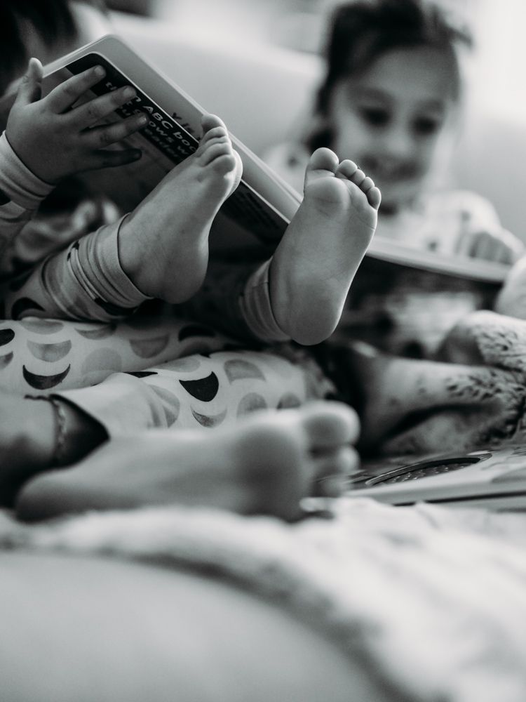 A black and white image shows small feet holding a mobile device while relaxing on patterned bedding.