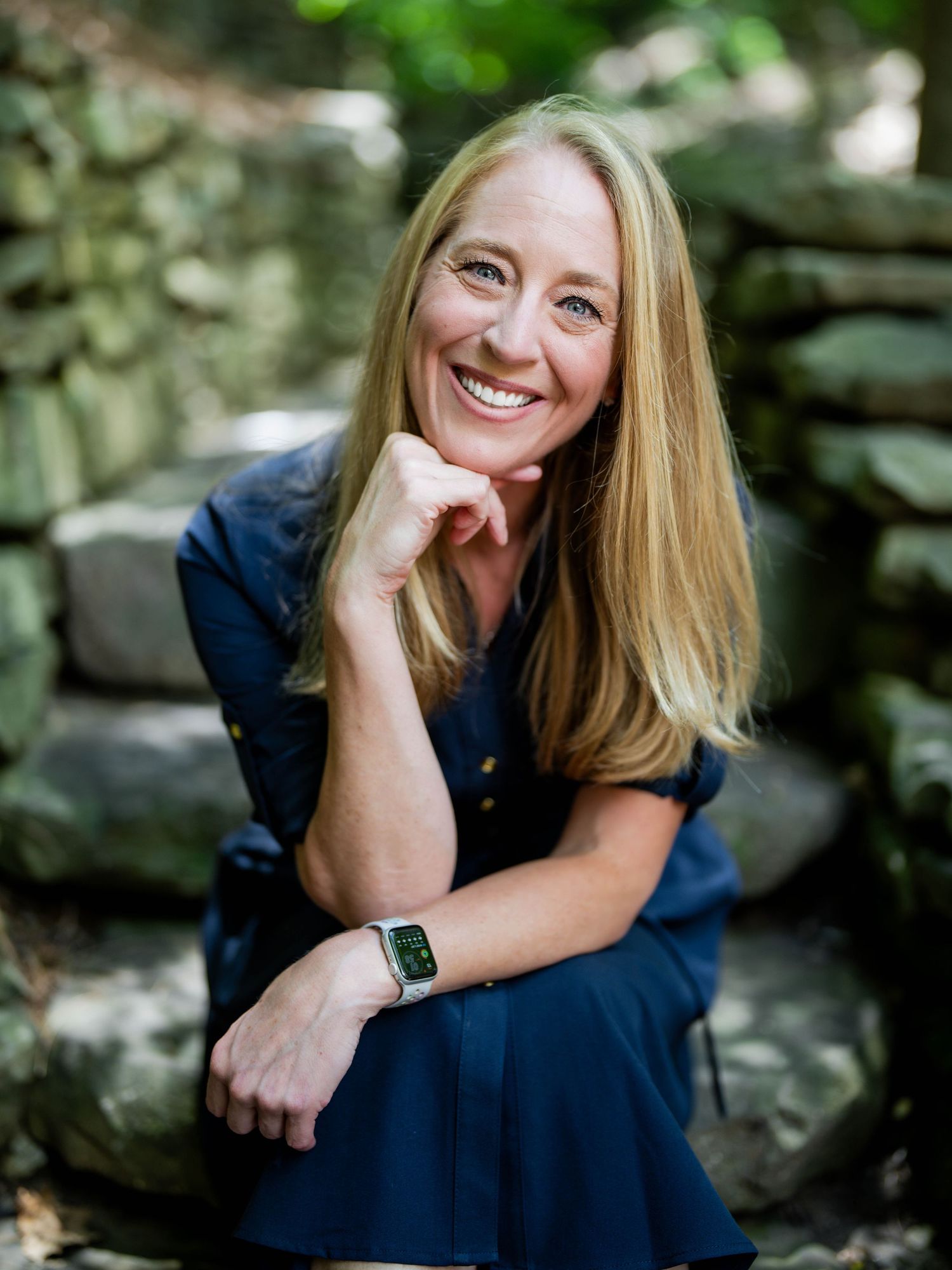Person in a navy blue dress sits on stone wall outdoors, resting chin on hand while smiling in natural setting.