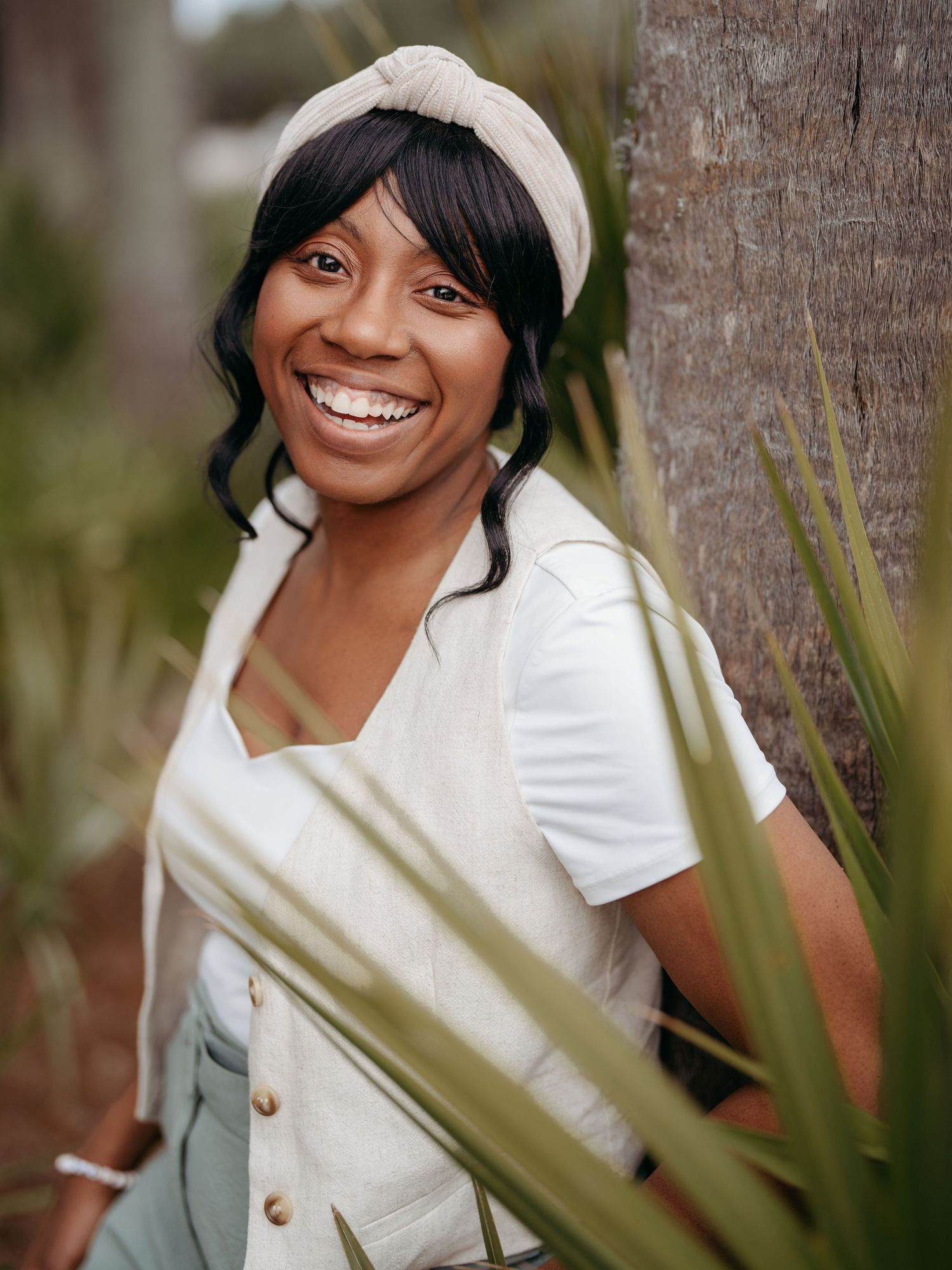 A bright smile radiates warmth as someone poses near a palm tree wearing a white top and cream headband.