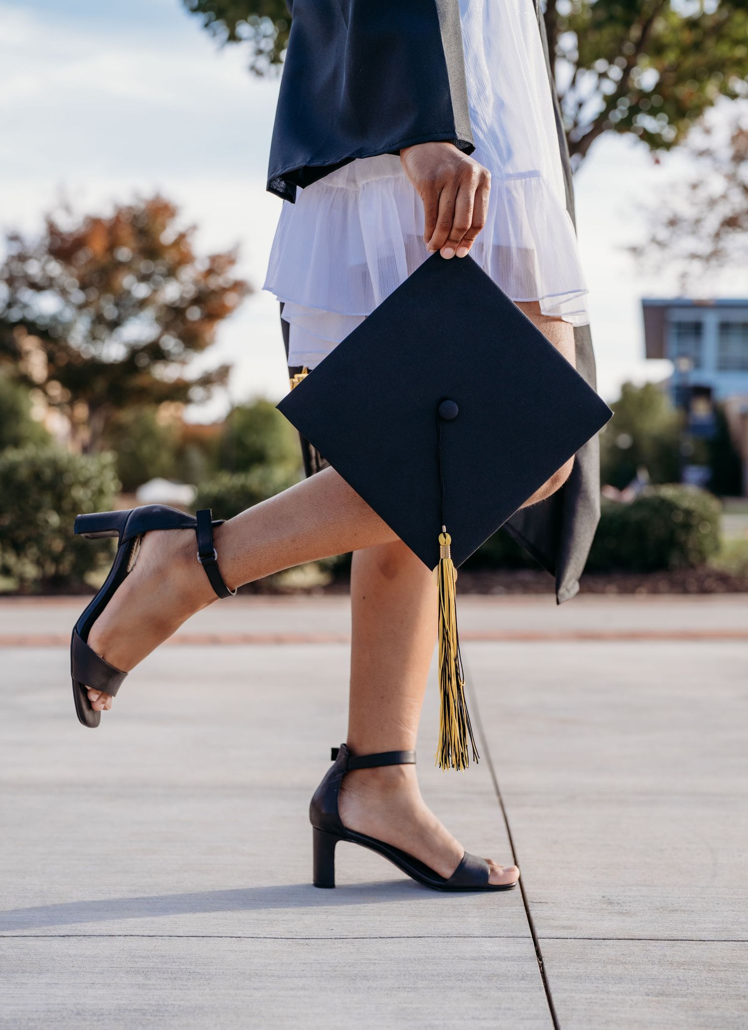 A graduate holding their black cap with gold tassel while wearing strappy heels and a white dress in an outdoor setting.