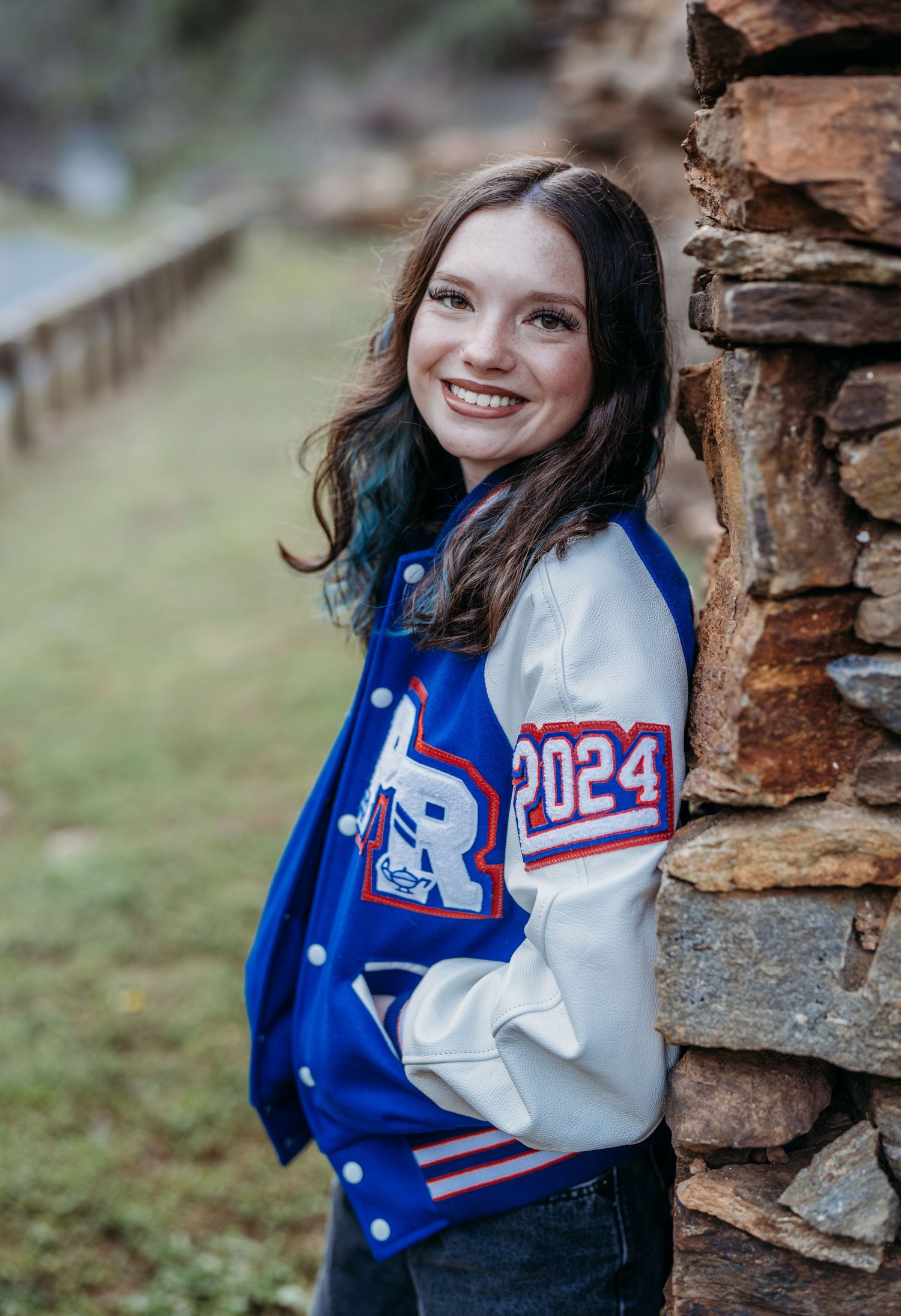 A varsity letterman jacket in blue and white with '2024' numbering is shown against a stone wall backdrop.