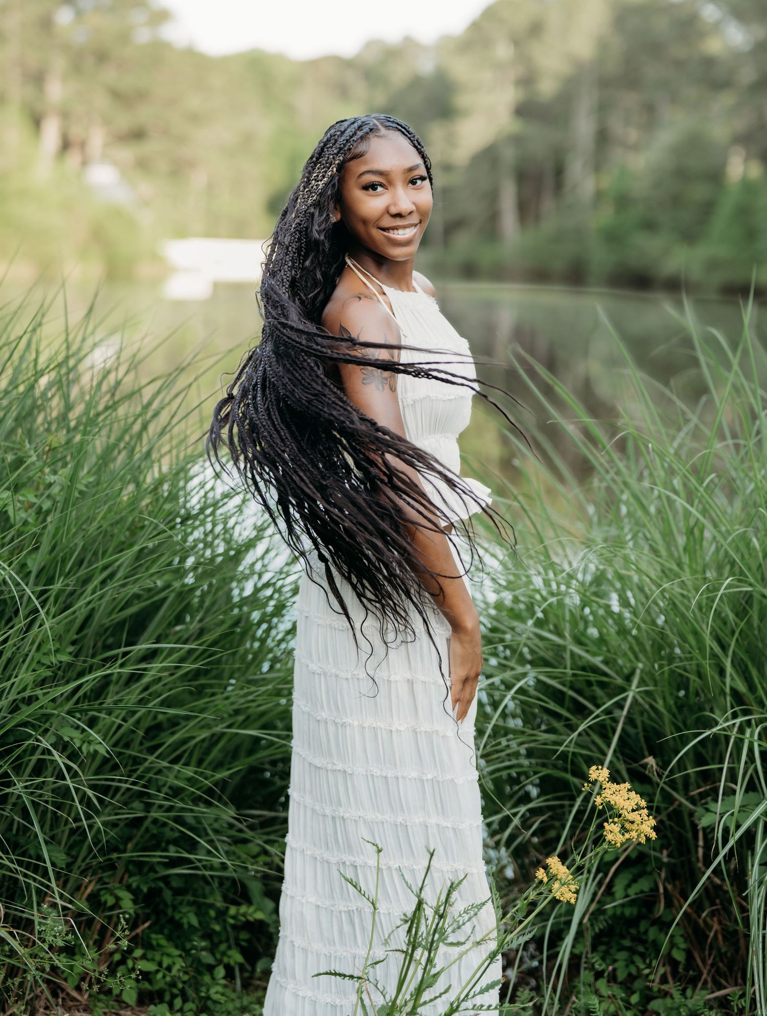 A person in a white dress poses gracefully among tall grasses by a lake during golden hour.