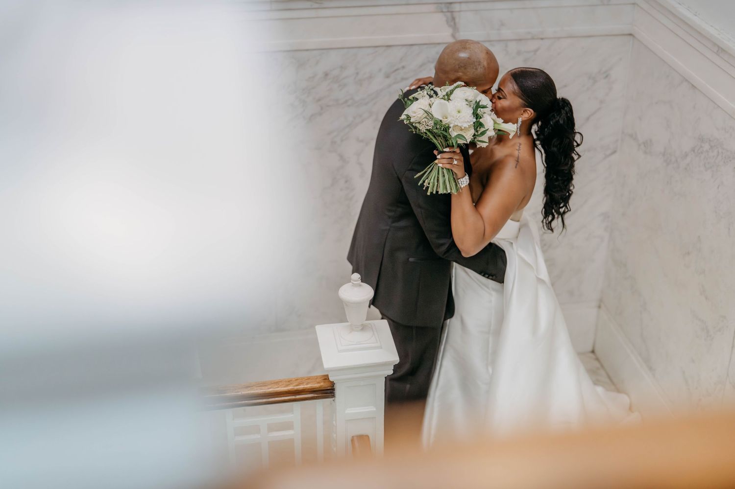 A romantic wedding couple shares a kiss in front of an elegant white marble wall during their special day.