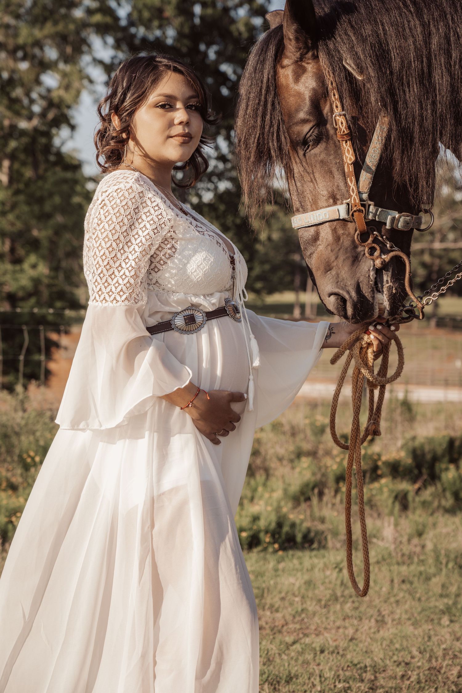 White lace maternity dress photography featuring a pregnant woman standing beside a brown horse in an outdoor setting.