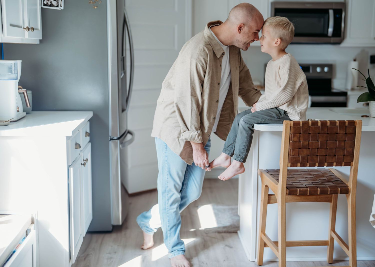 A Dad in beige jacket and jeans stands next to son, sharing a tender moment, with young boy perched on the kitchen countertop.
