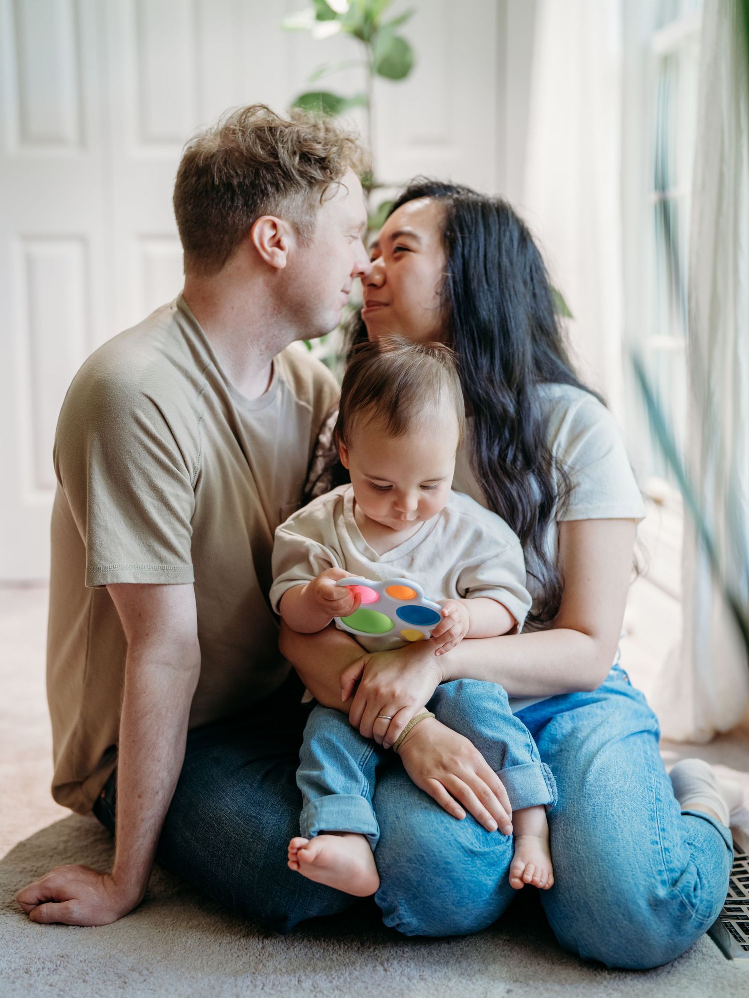 Couple sitting on floor with baby playing with colorful toy in bright room by window.