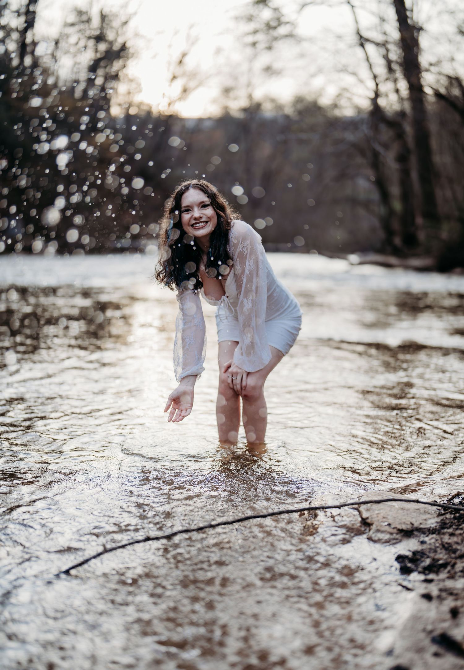 A joyful figure in a white dress splashes in a shallow stream during winter with snow falling in the forest.