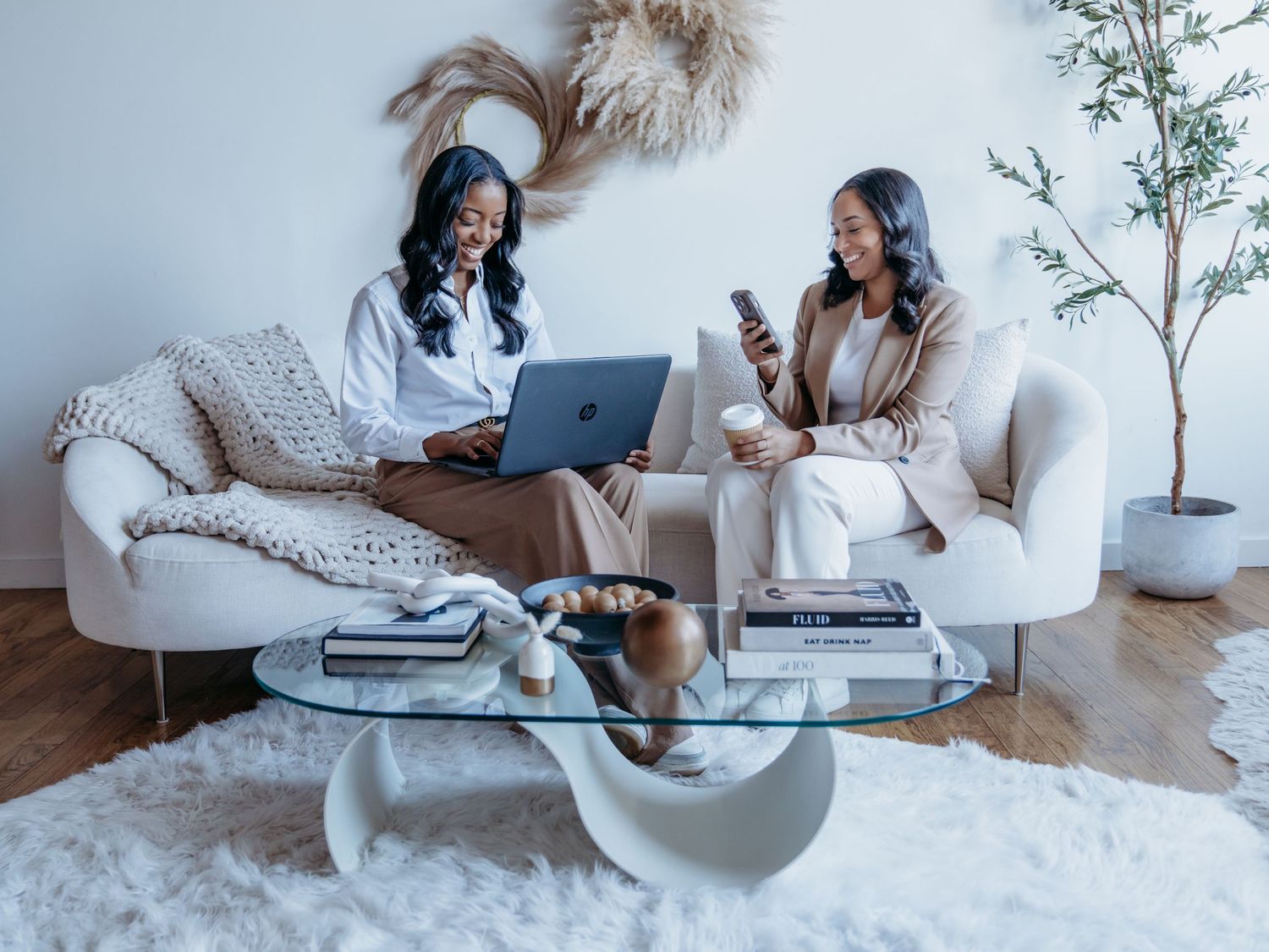 Two women sit on white armchairs having a business meeting with a laptop and coffee table in a bright modern room.