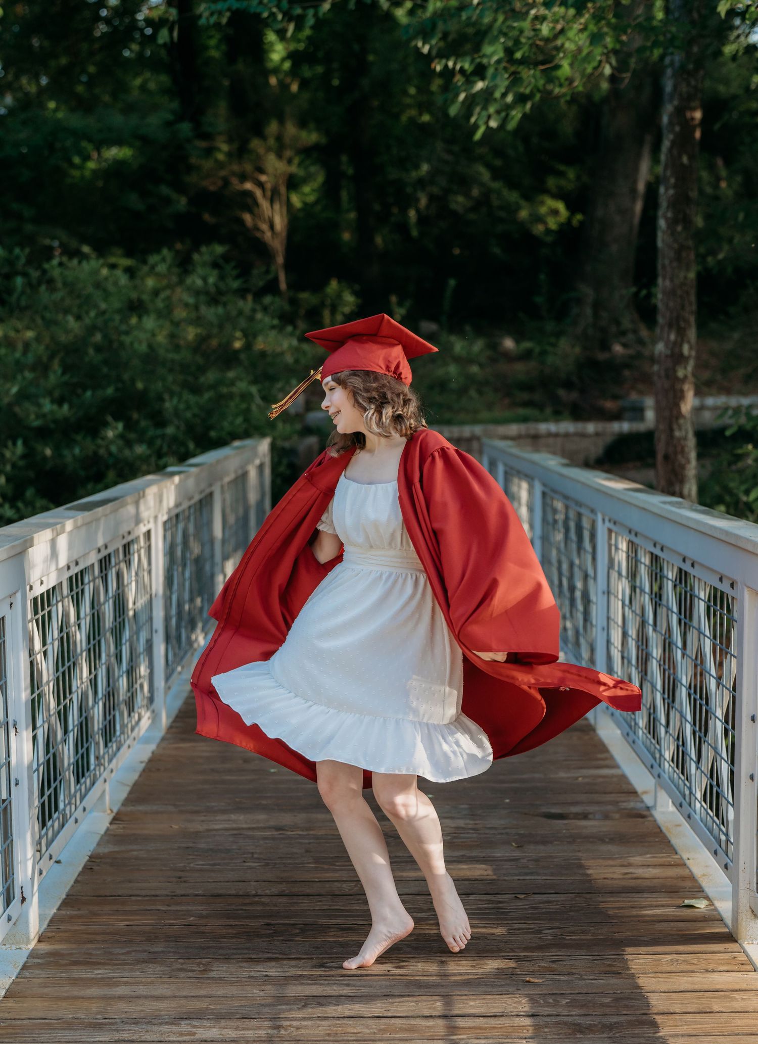 Person in white dress and flowing red graduation gown twirls gracefully on wooden bridge in forest setting.