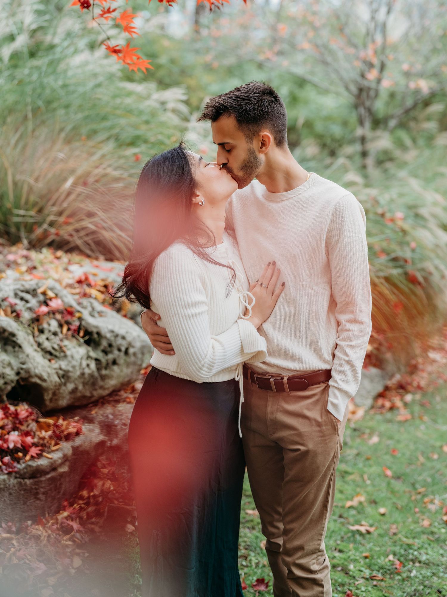 A romantic autumn garden photo session features a couple amid lush greenery and fallen red leaves.