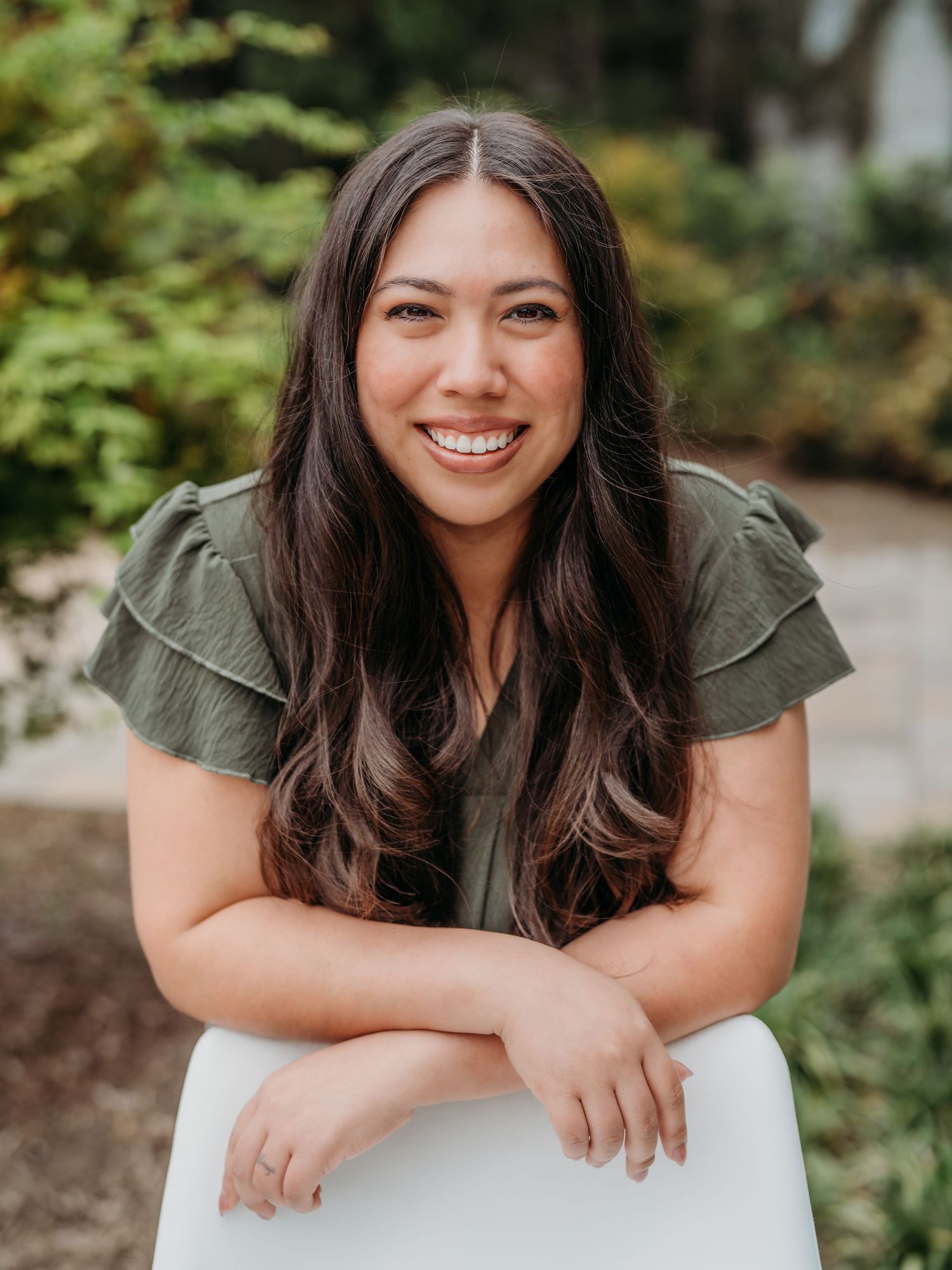 Woman in olive green blouse poses cheerfully against natural outdoor background.