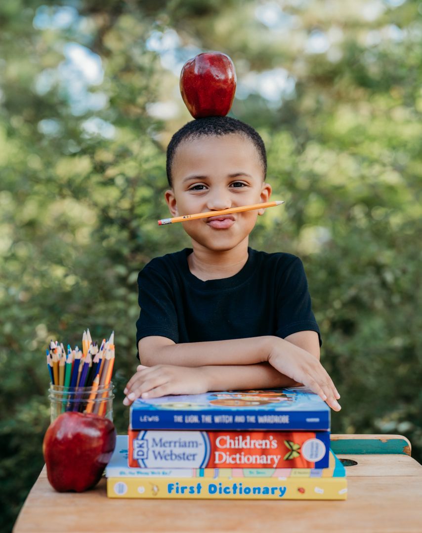 A smiling boy balances an apple on his head and pencil on his lip, sitting behind stacked dictionaries and colored pencils.