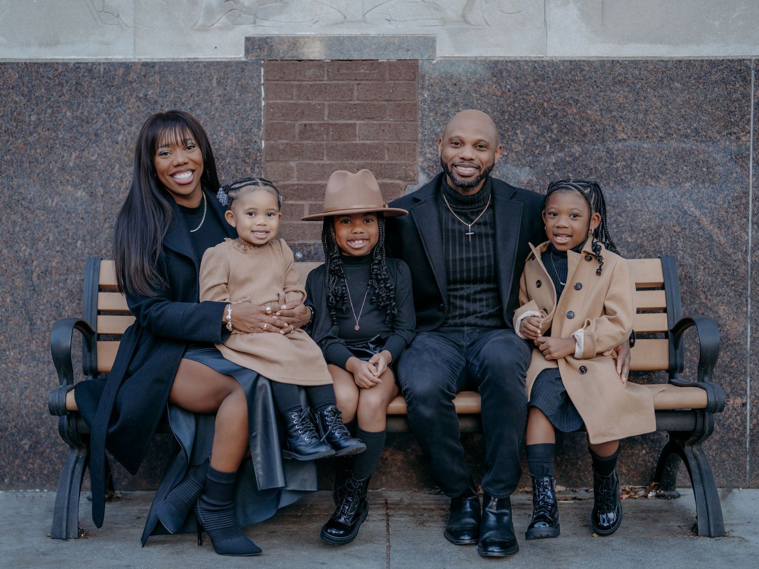 A family of five sits together on a wooden bench wearing coordinated winter outfits in dark and neutral tones.