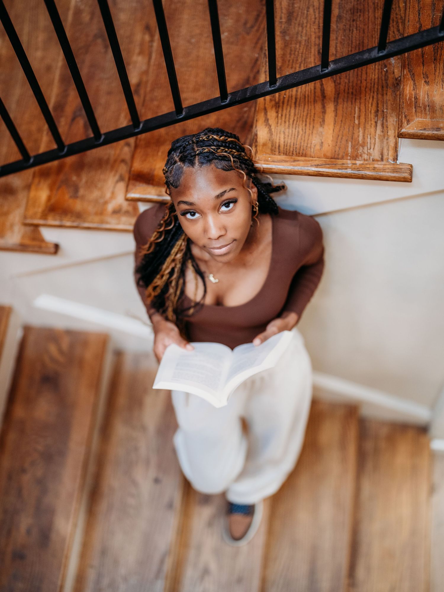 View from above of someone in a brown top and light pants sitting on wooden stairs while reading a book.