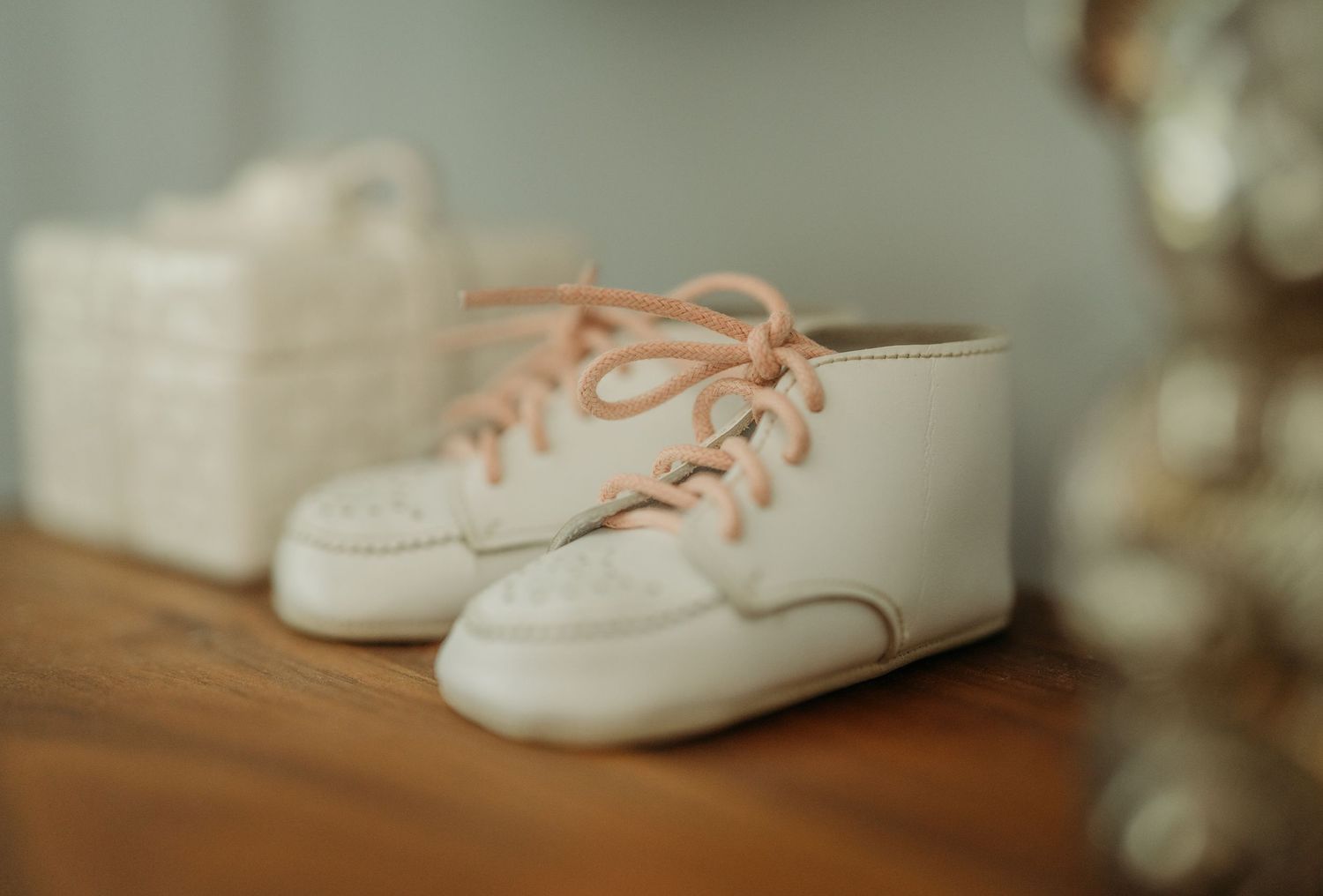 White leather baby moccasin shoes with coral laces sit on a wooden table in soft focus lighting.