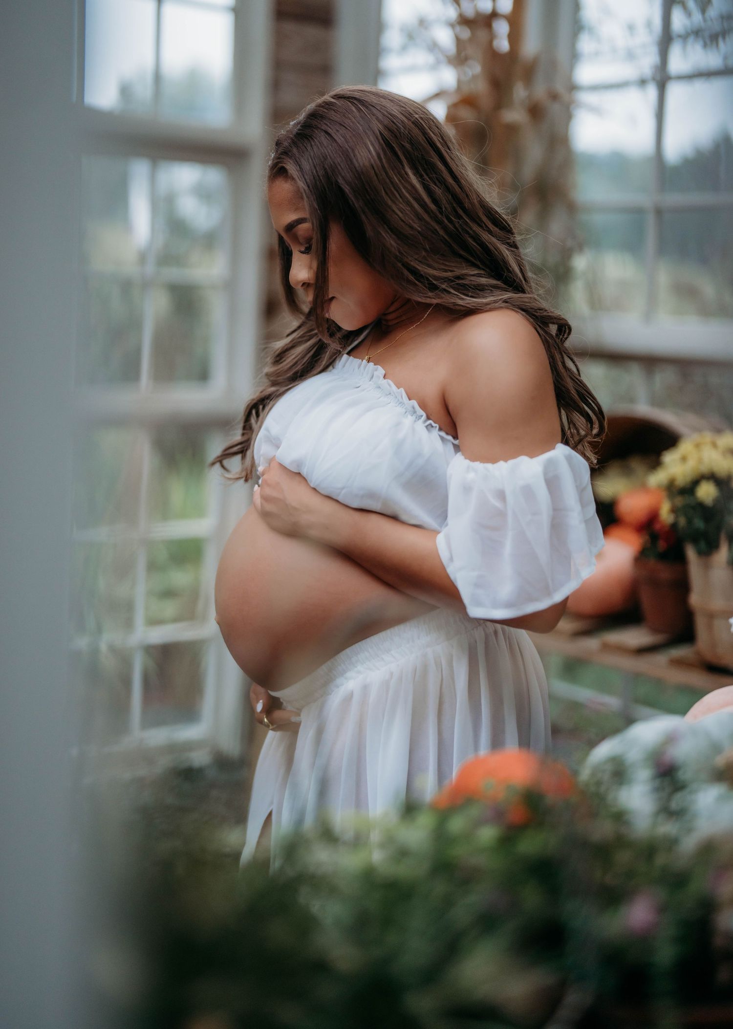 Expecting mother in white off-shoulder dress poses in sunlit greenhouse with flowers and plants.