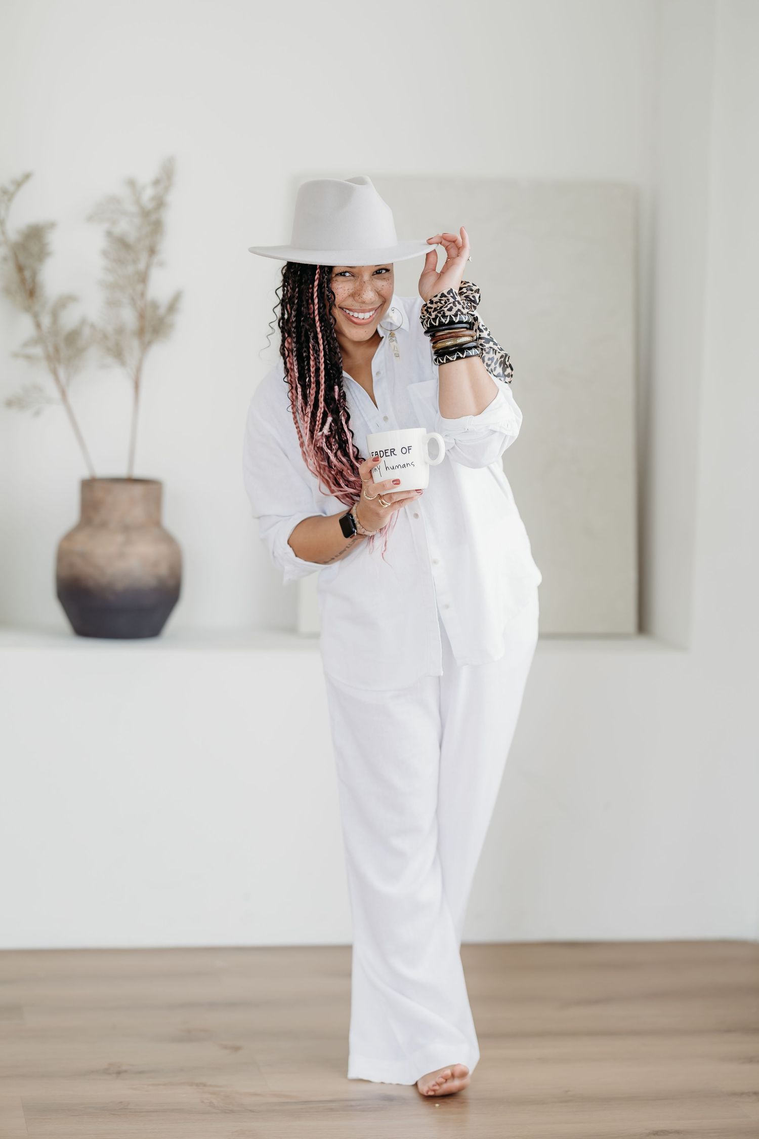A person in an all-white outfit with braids and stacked bracelets holds a coffee mug while standing in a minimal room.