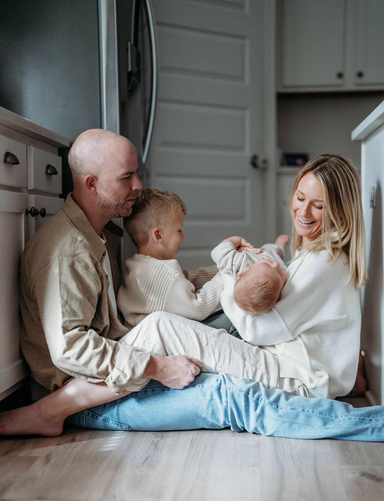 A family sits together on the kitchen floor in matching white sweaters, sharing an intimate moment at home.