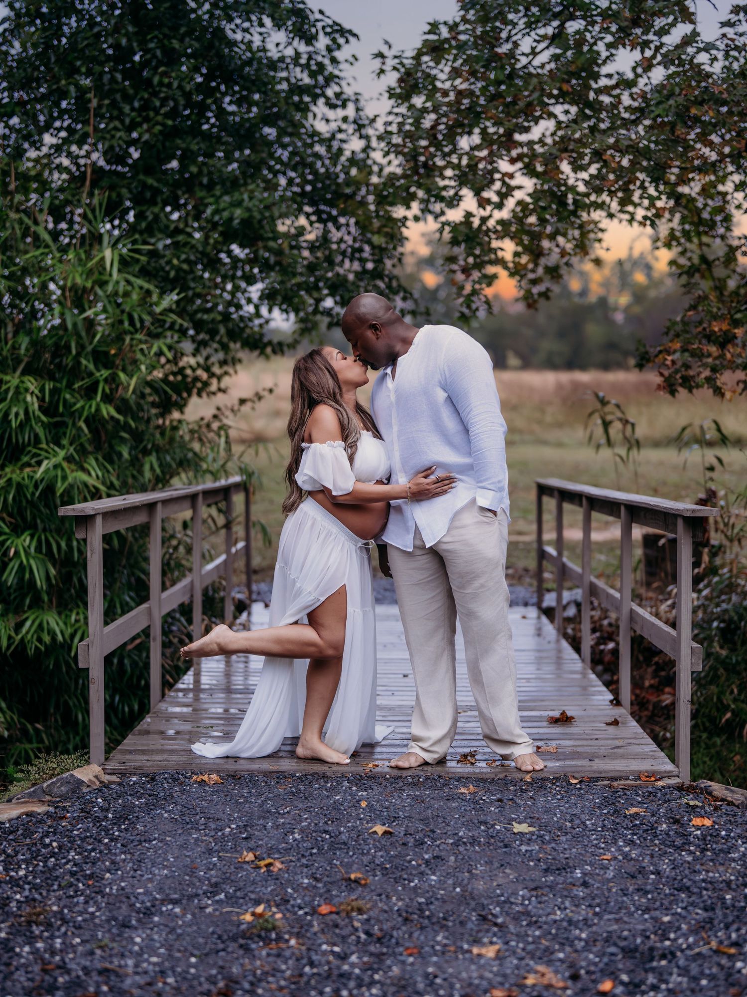 A romantic couple in white outfits embrace on a wooden bridge during sunset in a natural outdoor setting.