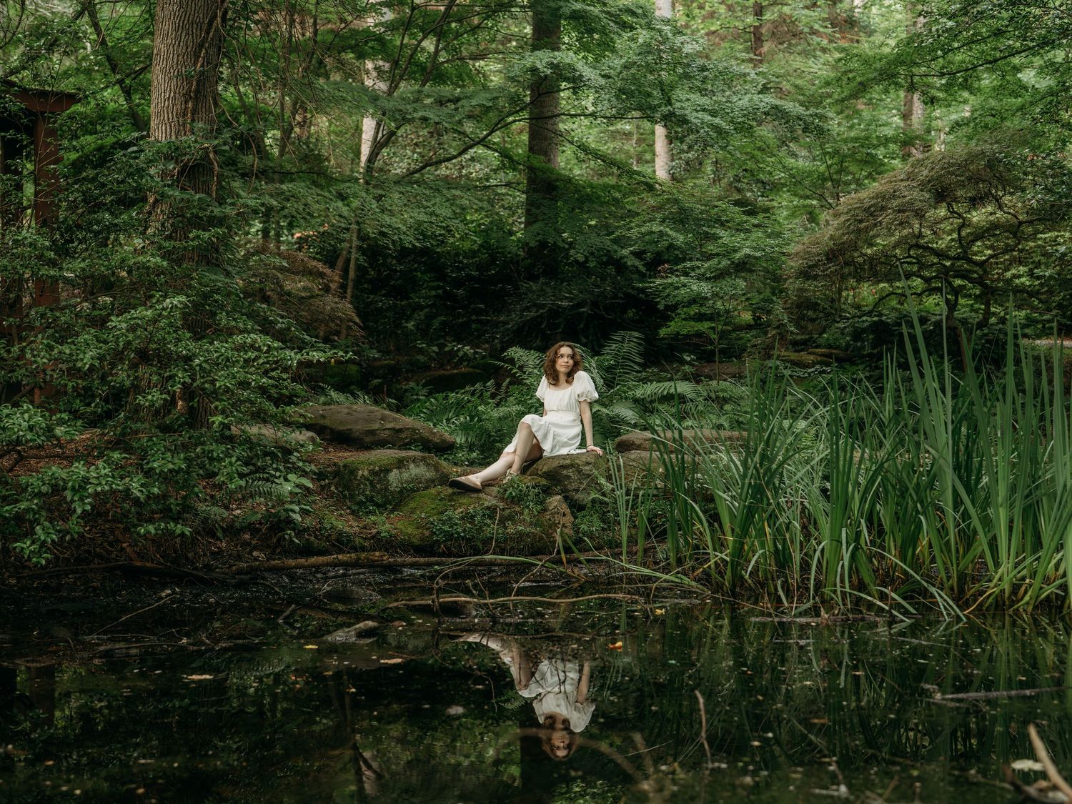 A serene forest pond reflects someone in a white dress sitting on a fallen log amidst lush greenery and cattails.