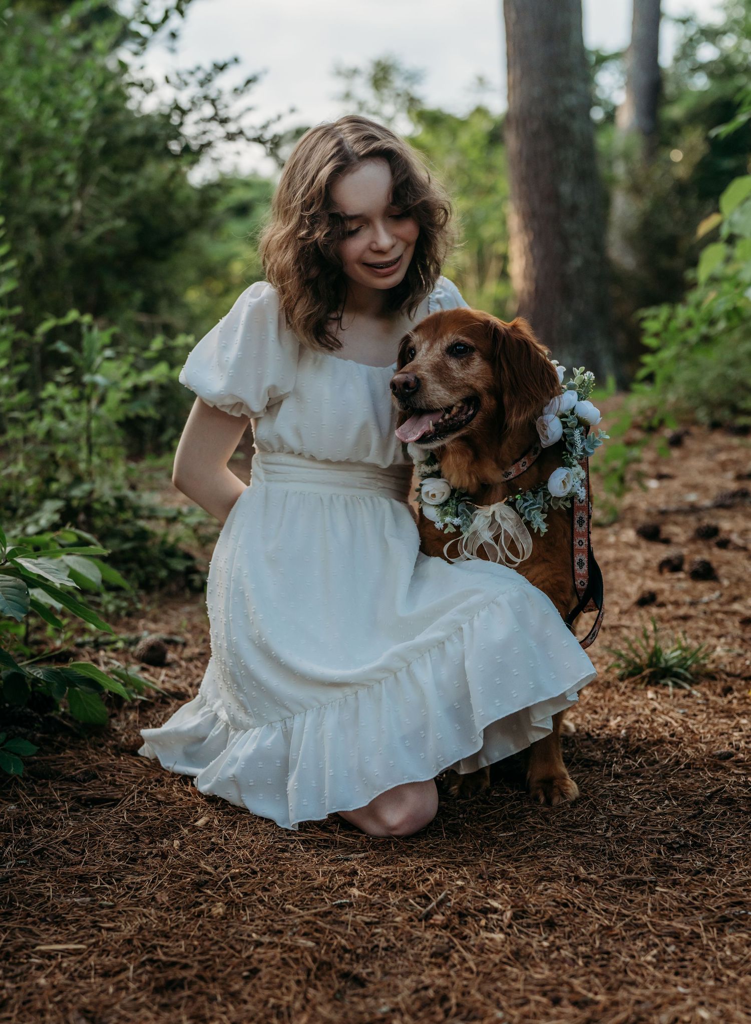 A brown dog wearing flowers sits on a forest path with someone in a flowing white dress kneeling beside them.