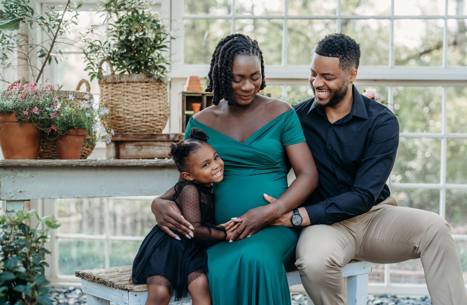 Expectant family shares a tender moment together in a sunlit room with potted plants.