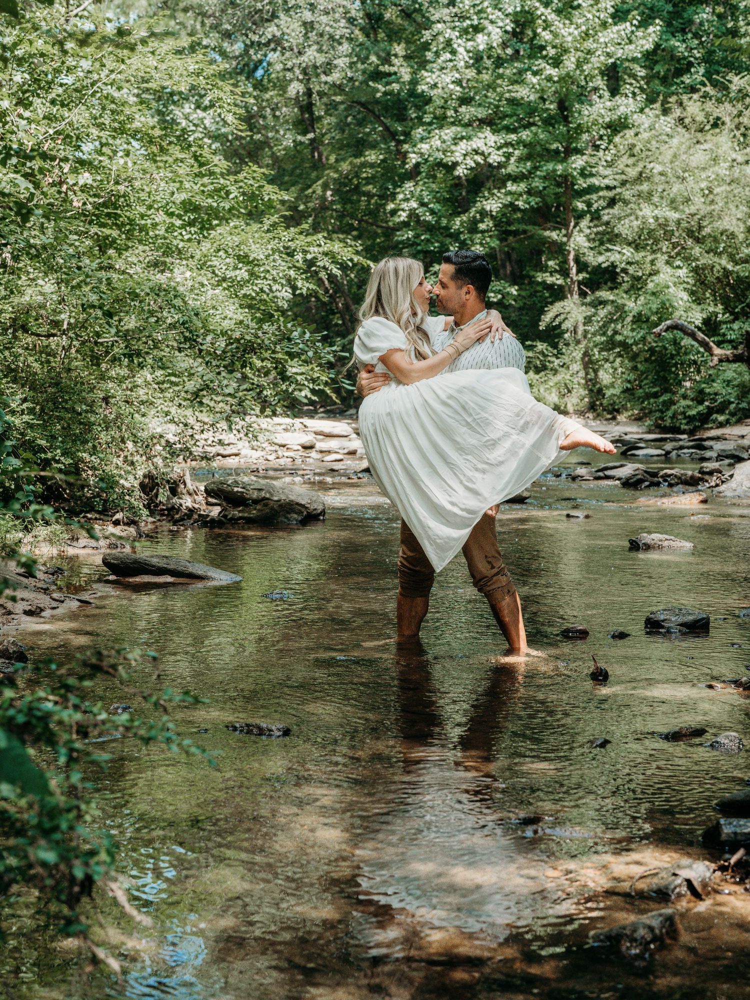A couple in white clothing shares a romantic moment while standing in a shallow creek surrounded by lush greenery.