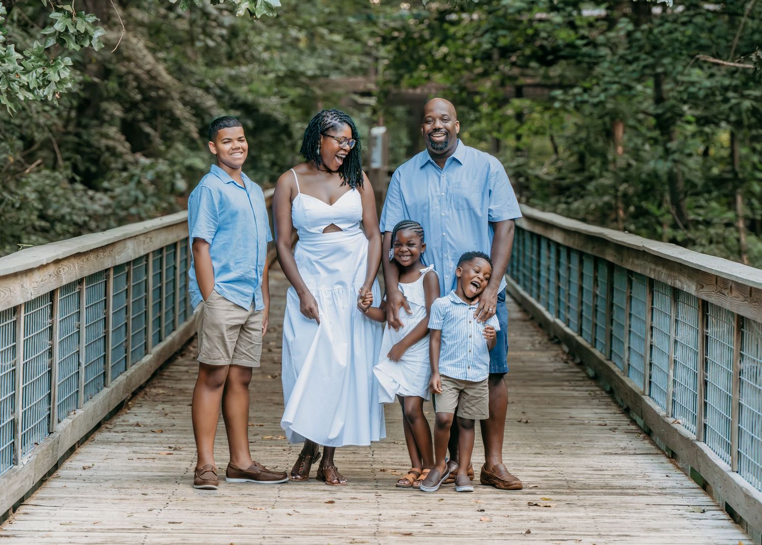A group in coordinating light blue and white outfits poses together on a wooden bridge in a forest setting.