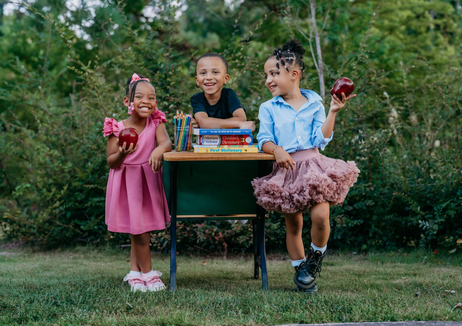 Three happy children sit at a school desk outdoors holding apples, surrounded by books and pencils in a lush green garden.
