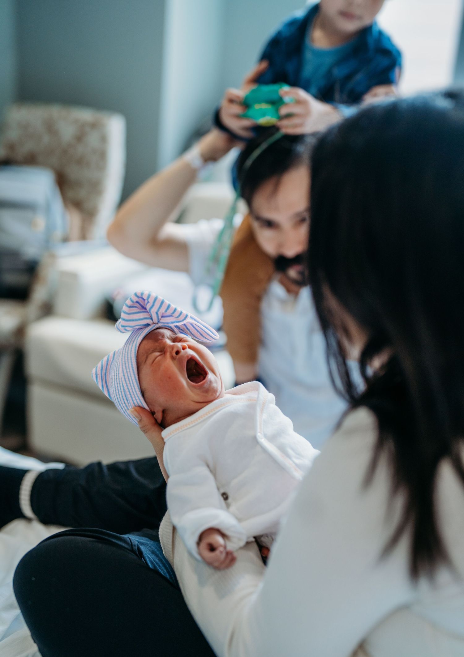A newborn baby in white clothing and small hat yawns while lying down as others gather around during a family moment.