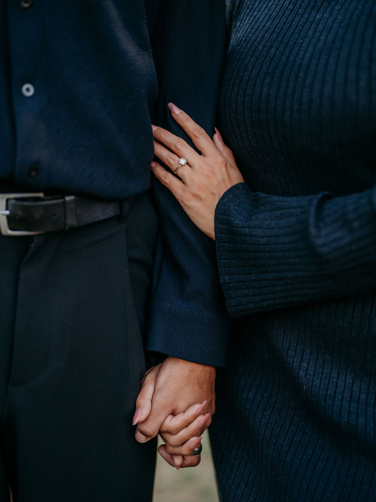Close-up shot of hands holding each other wearing navy blue sweater and suit showing an engagement ring.