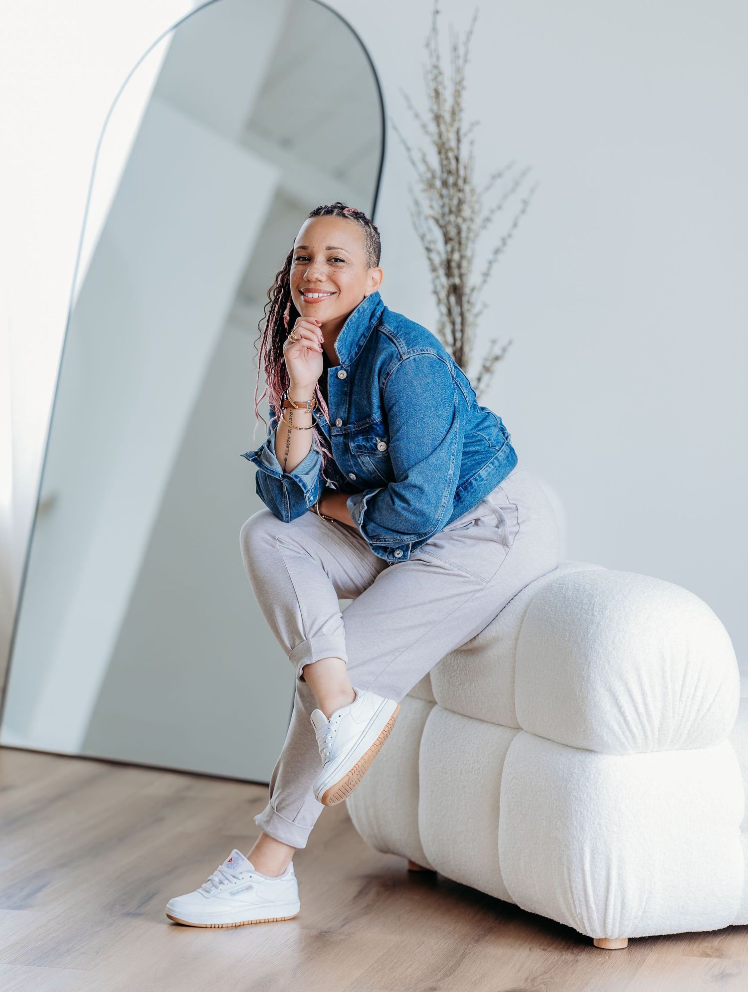 A person in a denim jacket sits casually on a white modular sofa against a light wall with decorative dry plants.