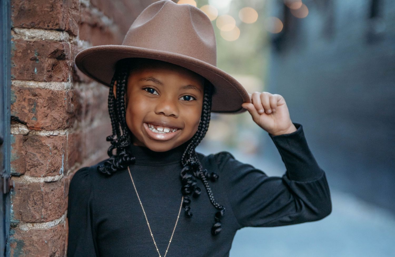 A person in a black turtleneck sweater and hat poses against a brick wall with a blurred urban background.
