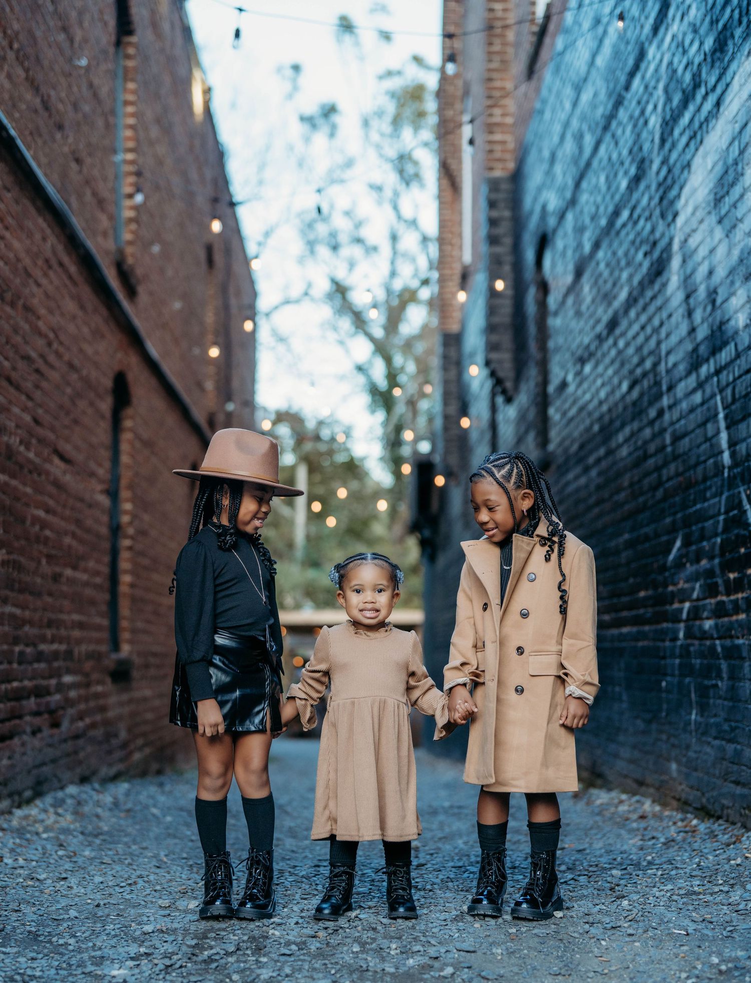Three young girls in contrasting black and beige outfits stand together in a brick alley.