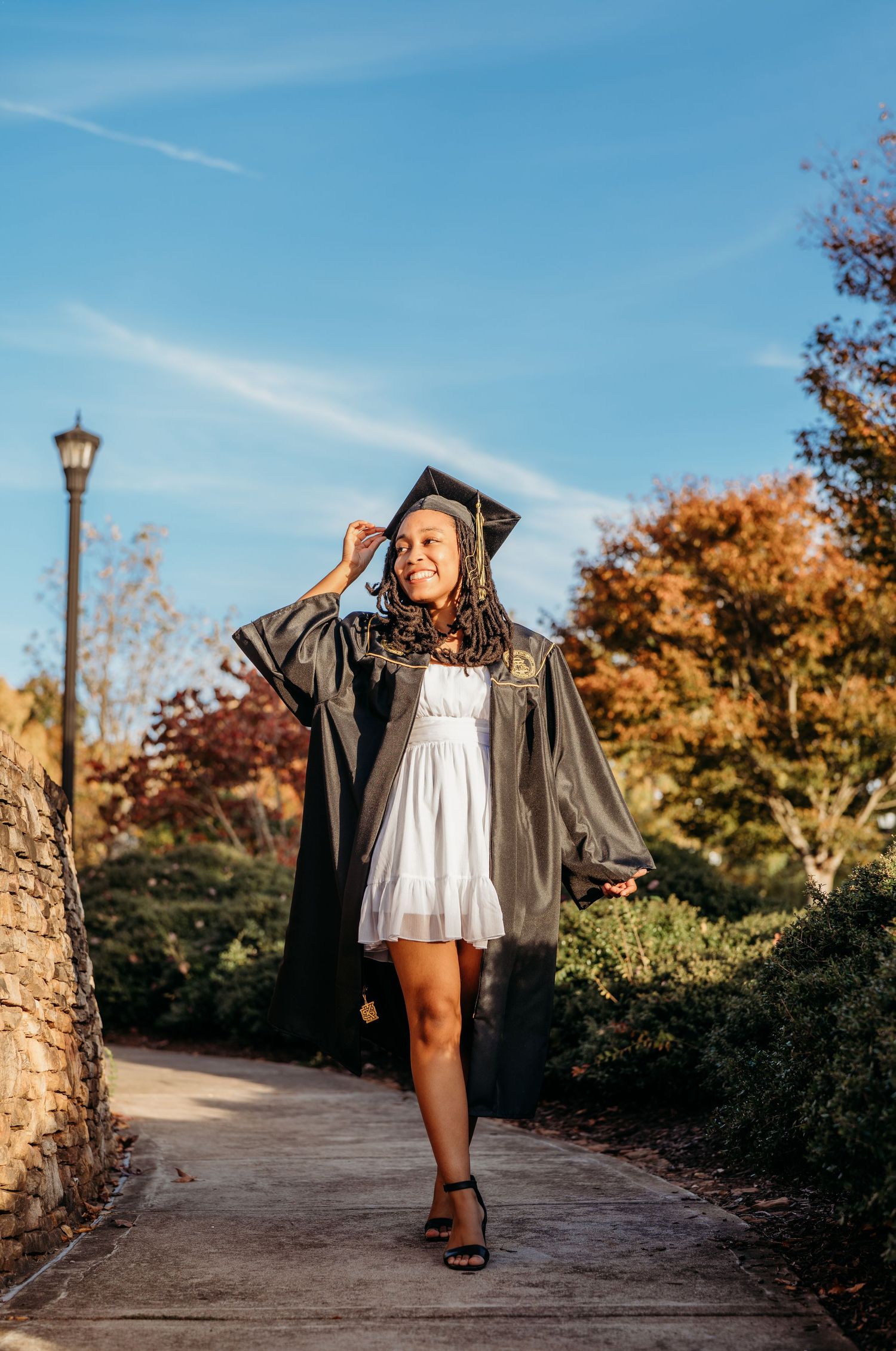 Graduate in white dress and black graduation gown poses on stone pathway during autumn with fall foliage and blue sky.