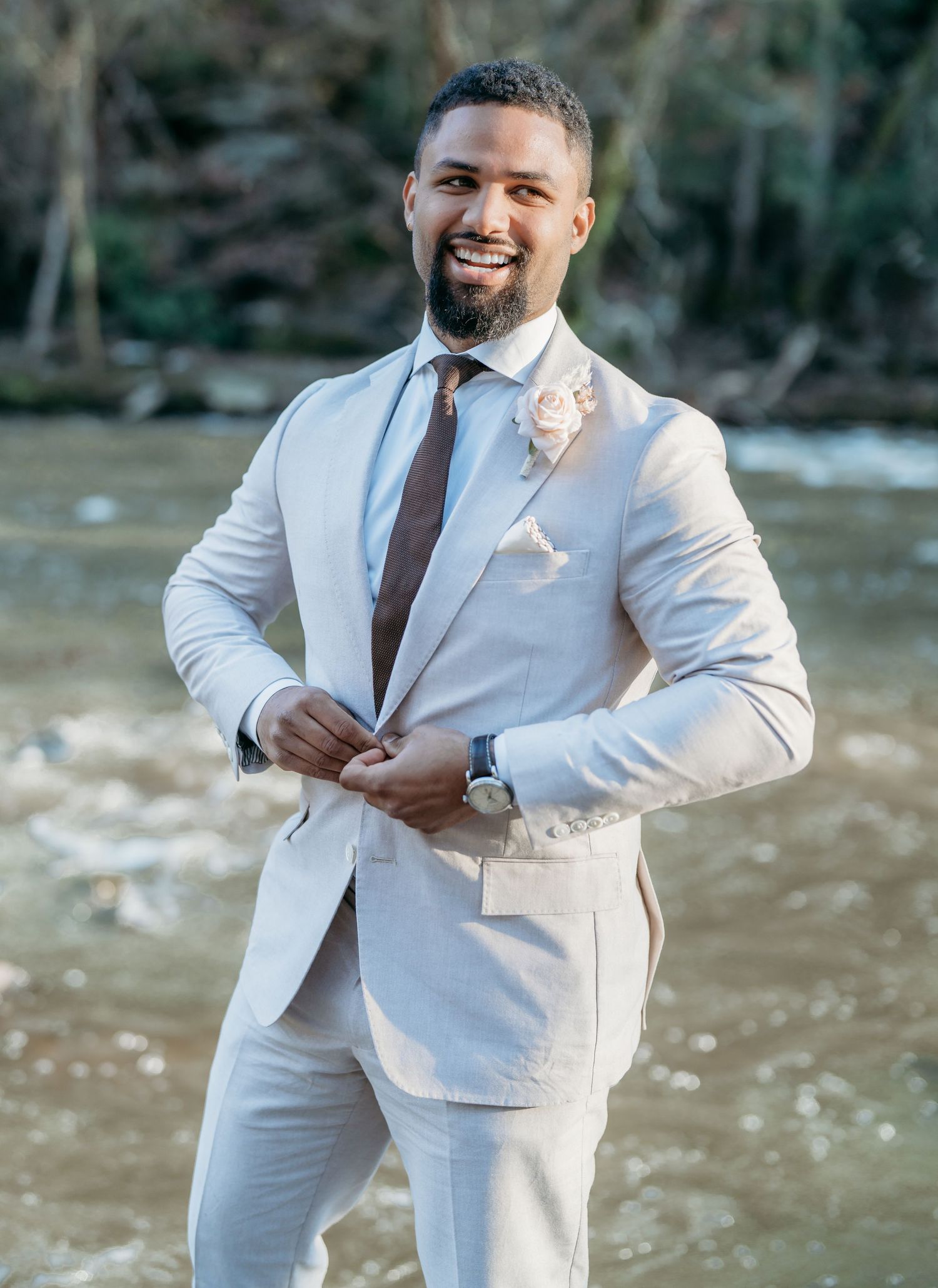 A groom in a light beige suit and brown tie poses outdoors near water with a boutonniere on his lapel.