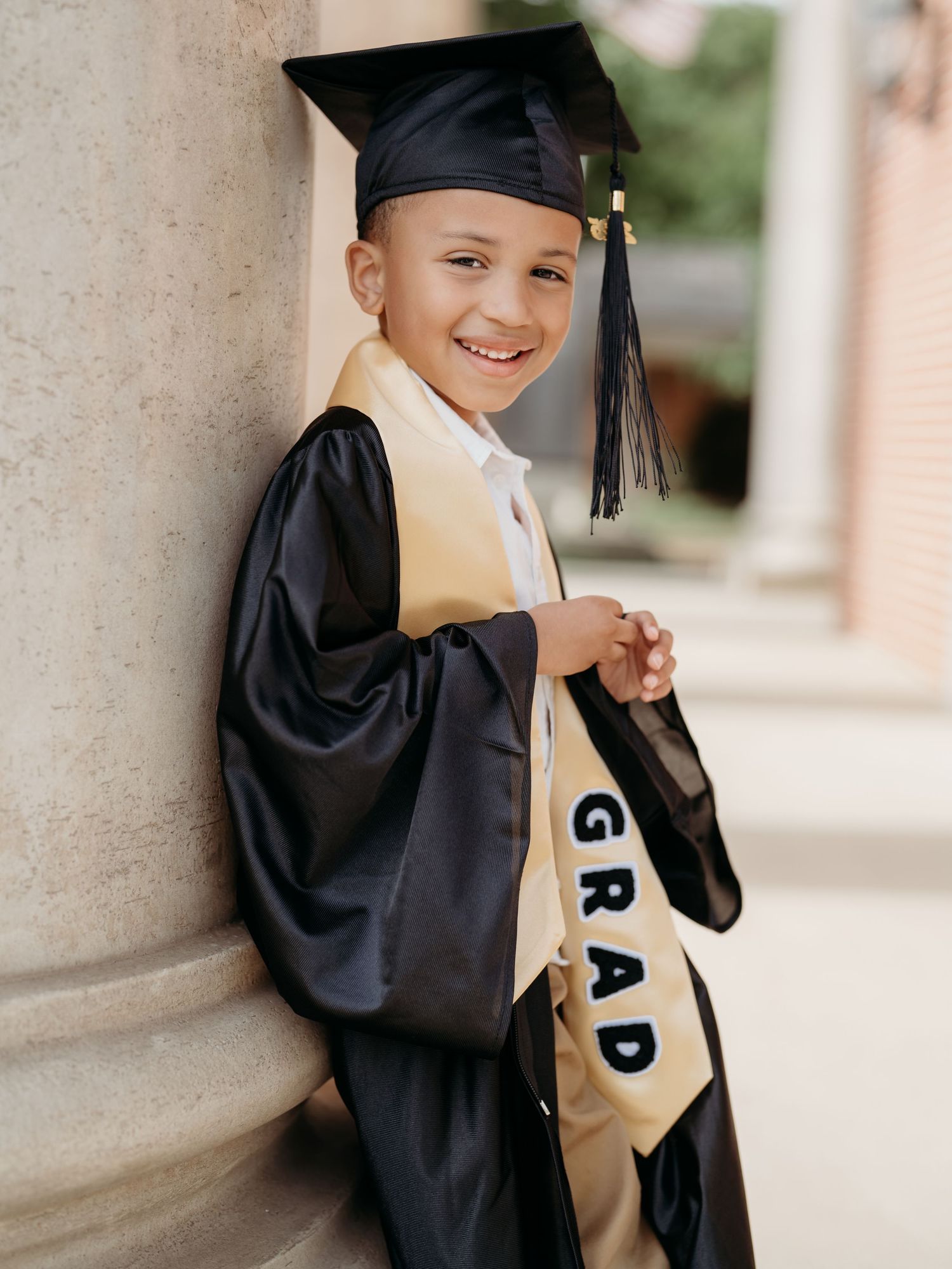 Young child smiling in black graduation cap and gown with gold GRAD sash, leaning against a column.