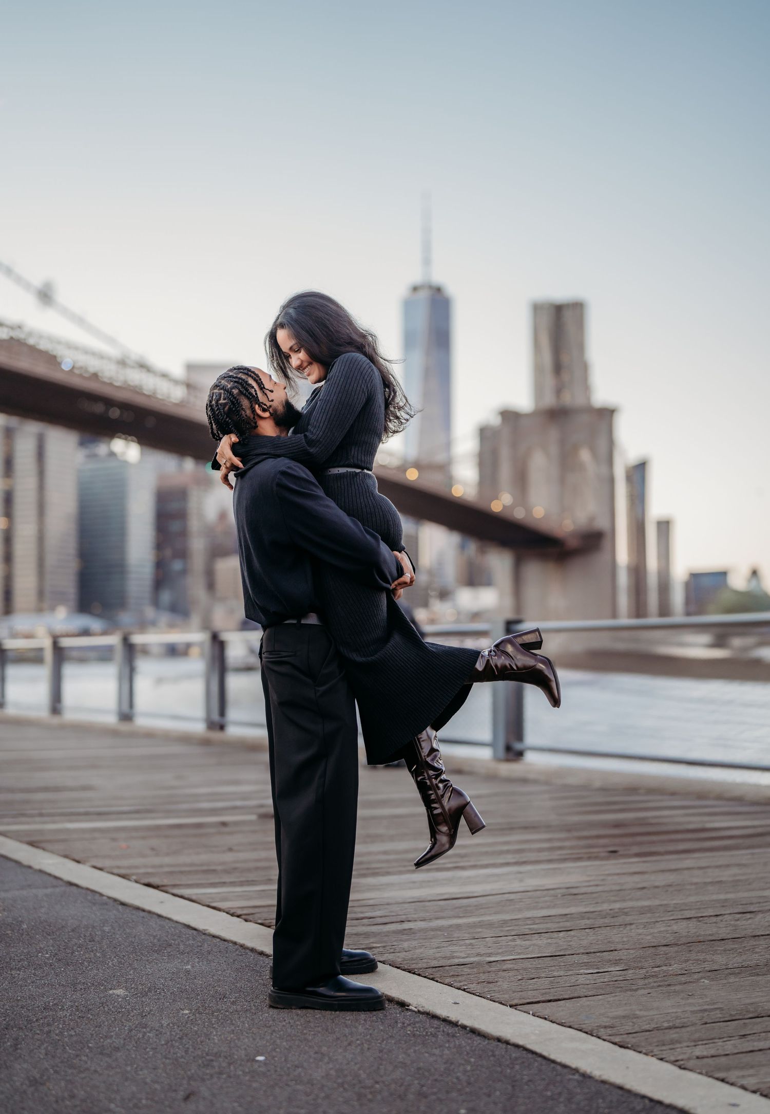 A romantic embrace on a waterfront boardwalk with the New York City skyline in the background.
