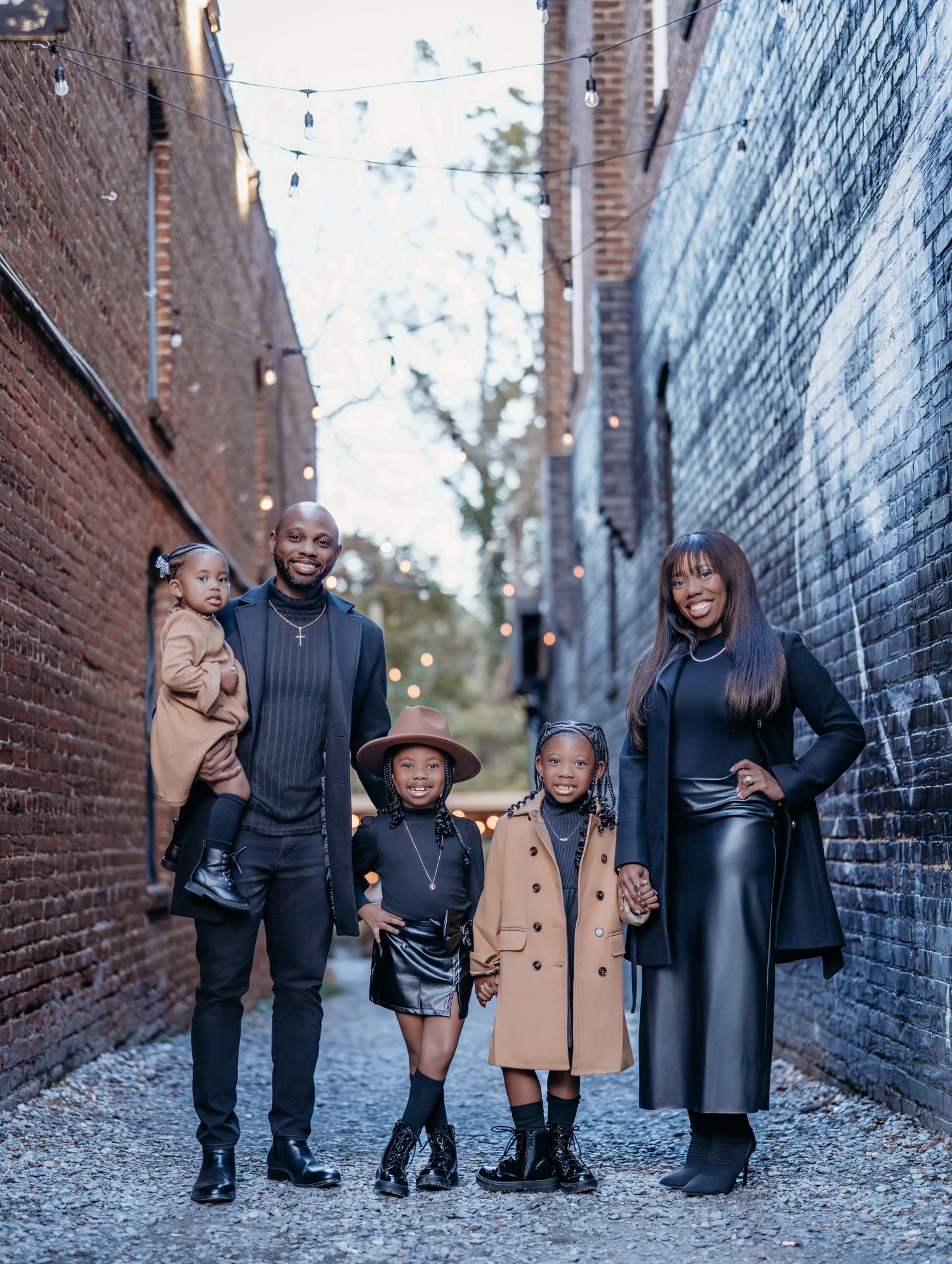 A family in coordinated black and tan outfits poses together in an urban alleyway with brick walls.