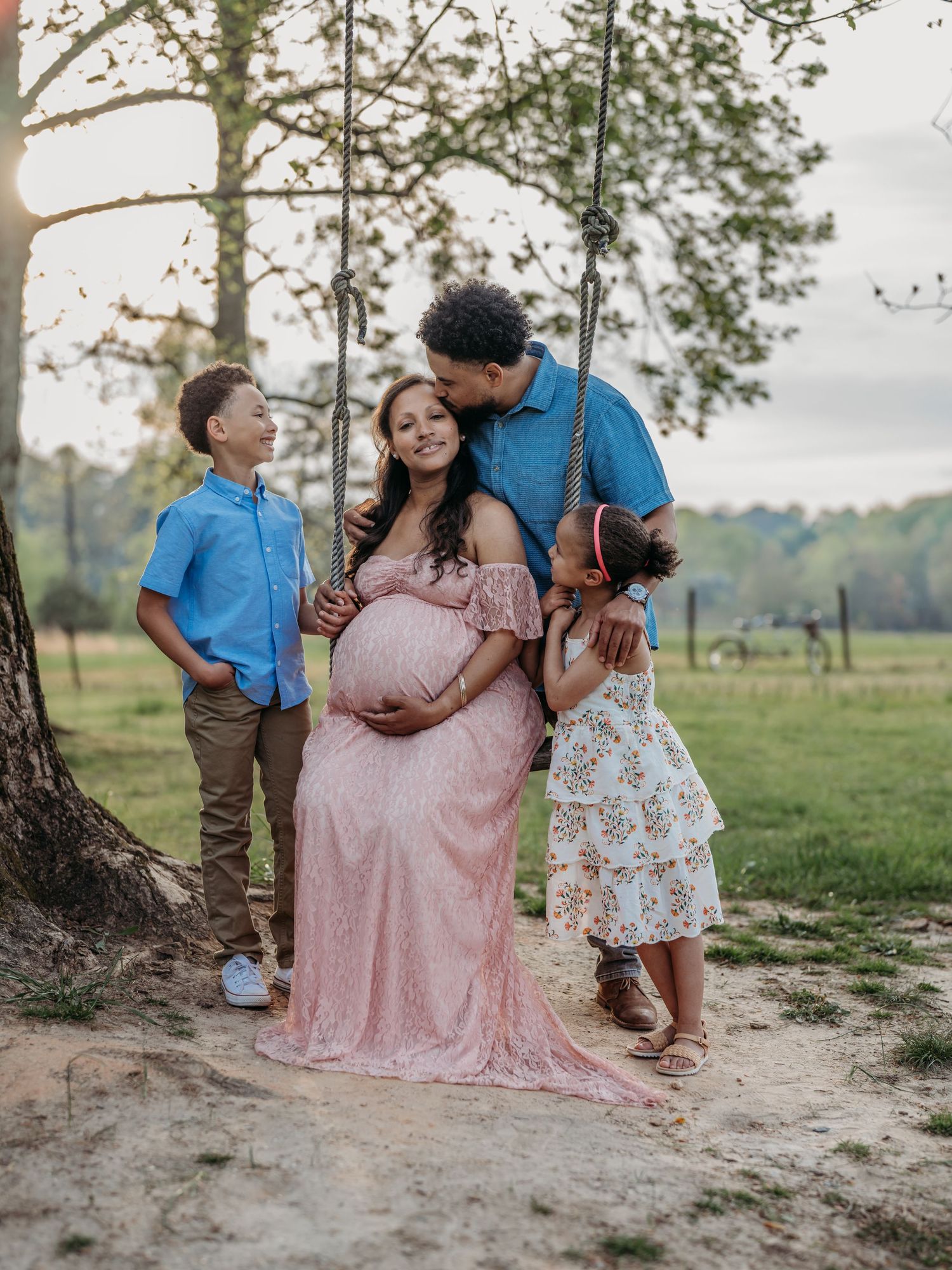 A family shares a tender moment outdoors in a pastoral setting with a pregnant woman in a pink lace gown.