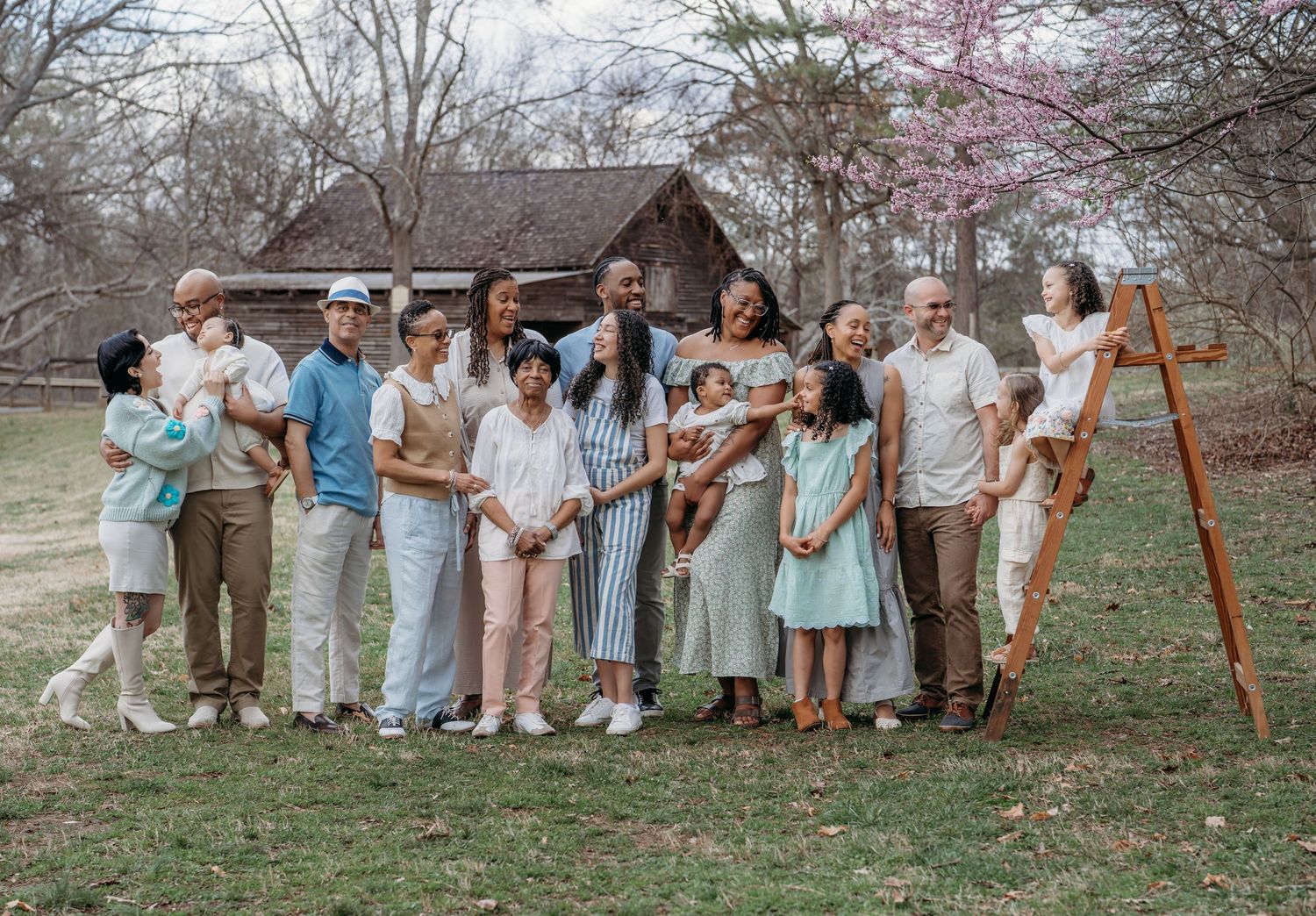 Group portrait outdoors in muted pastel clothing standing together on grassy field with blooming trees in background.