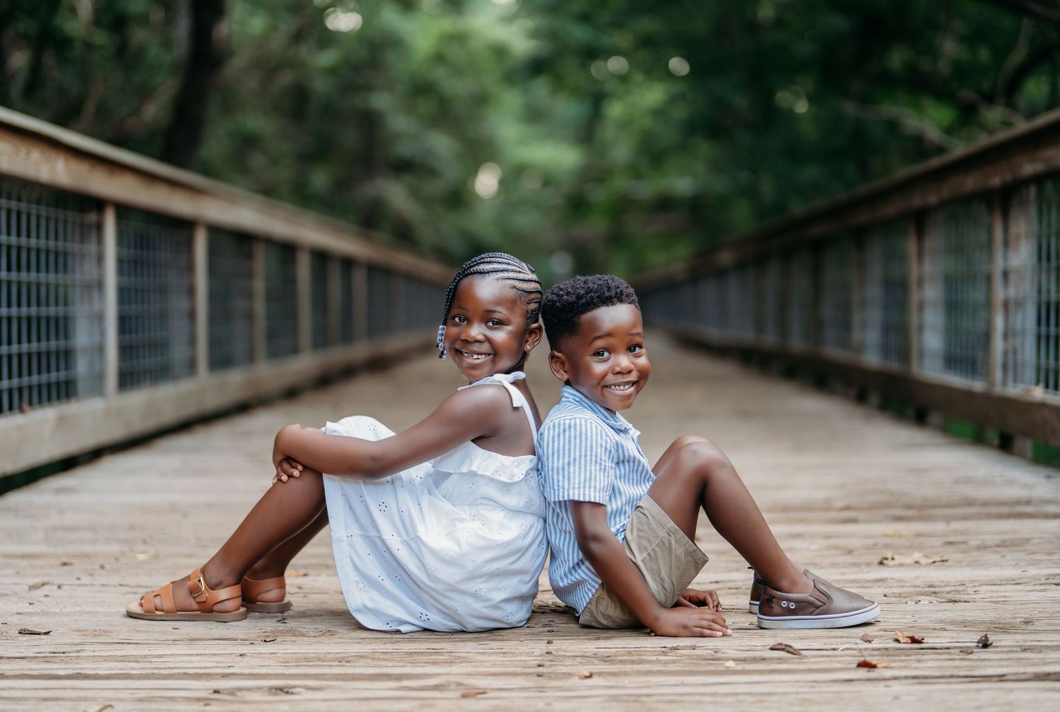 Two children lay playfully on a wooden bridge or walkway in matching light-colored outfits.
