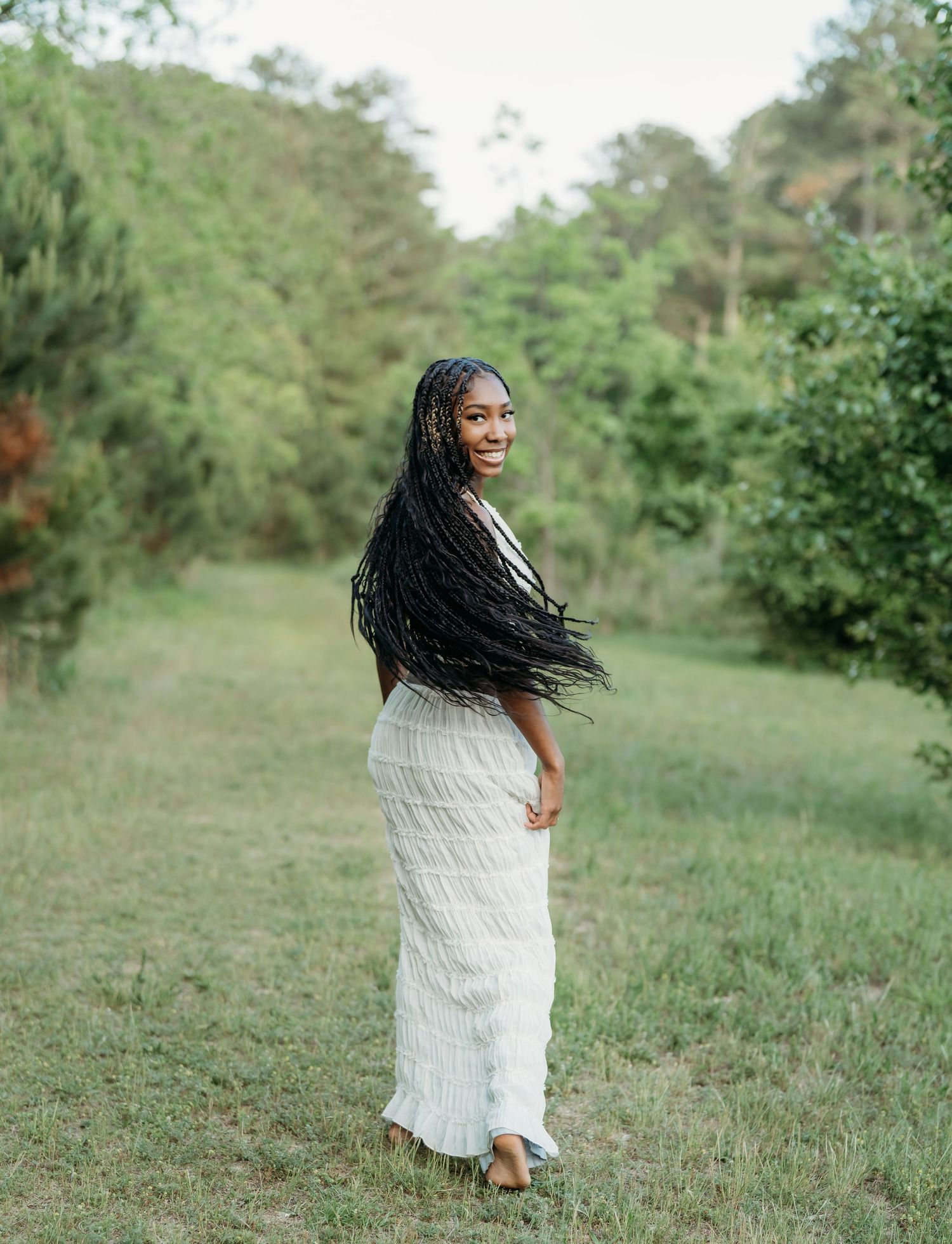 A figure in a long white dress stands in a grassy outdoor setting surrounded by lush greenery and trees.