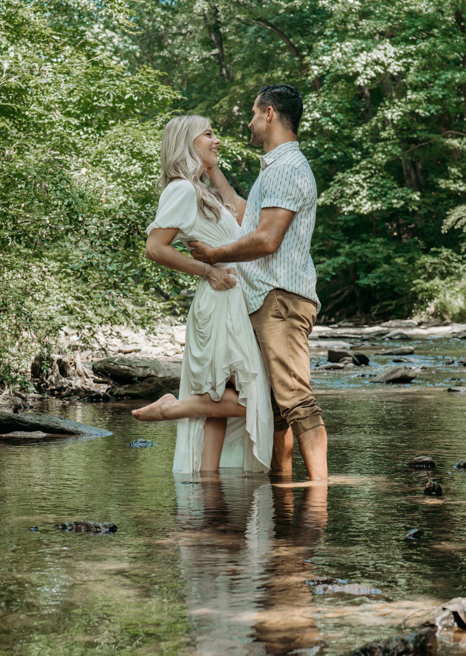 A couple shares a romantic moment while standing in a shallow creek surrounded by lush greenery on a summer day.