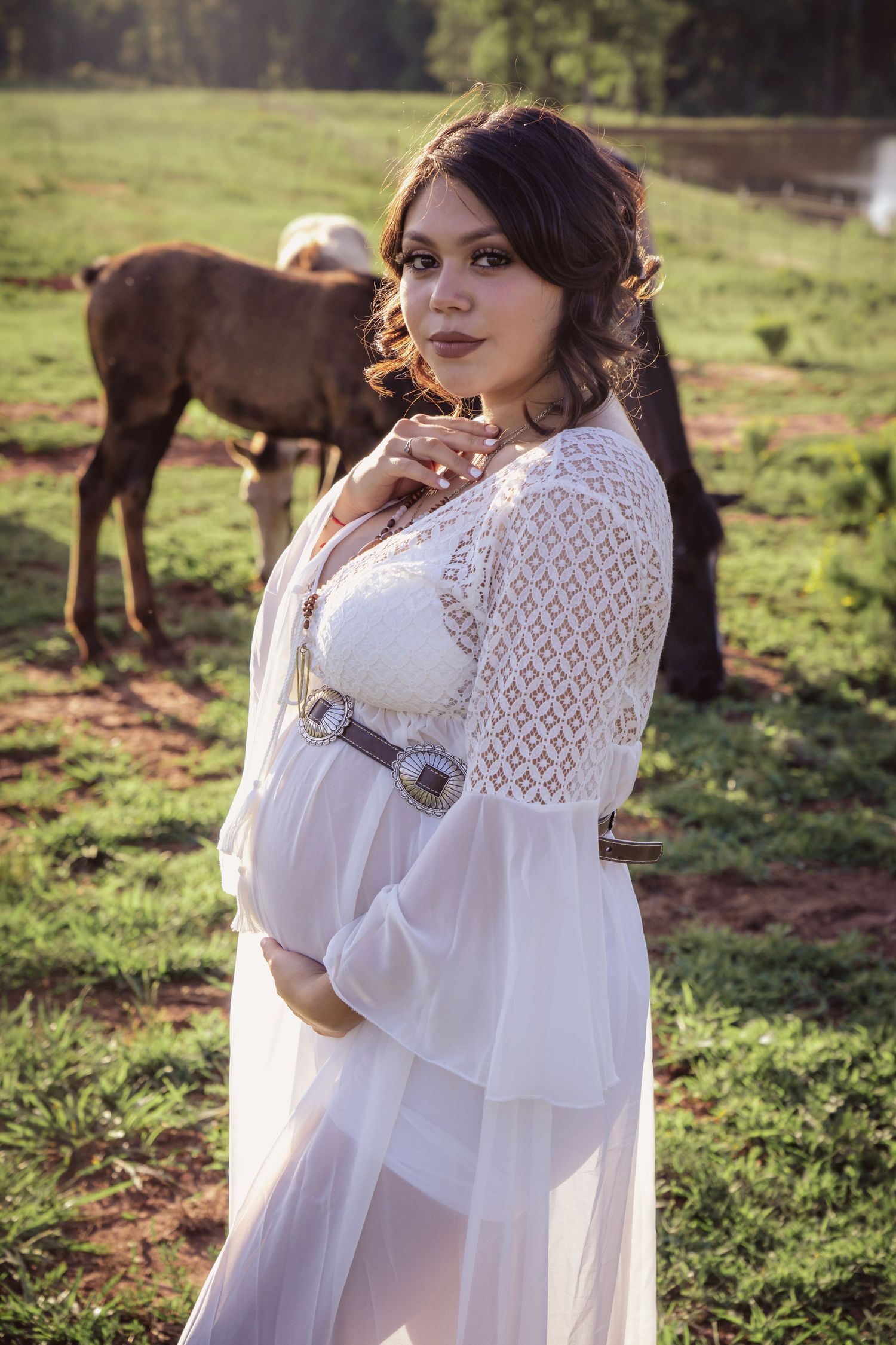 A woman in a flowing white dress and lace shawl poses in a sunlit field with horses grazing in the background.